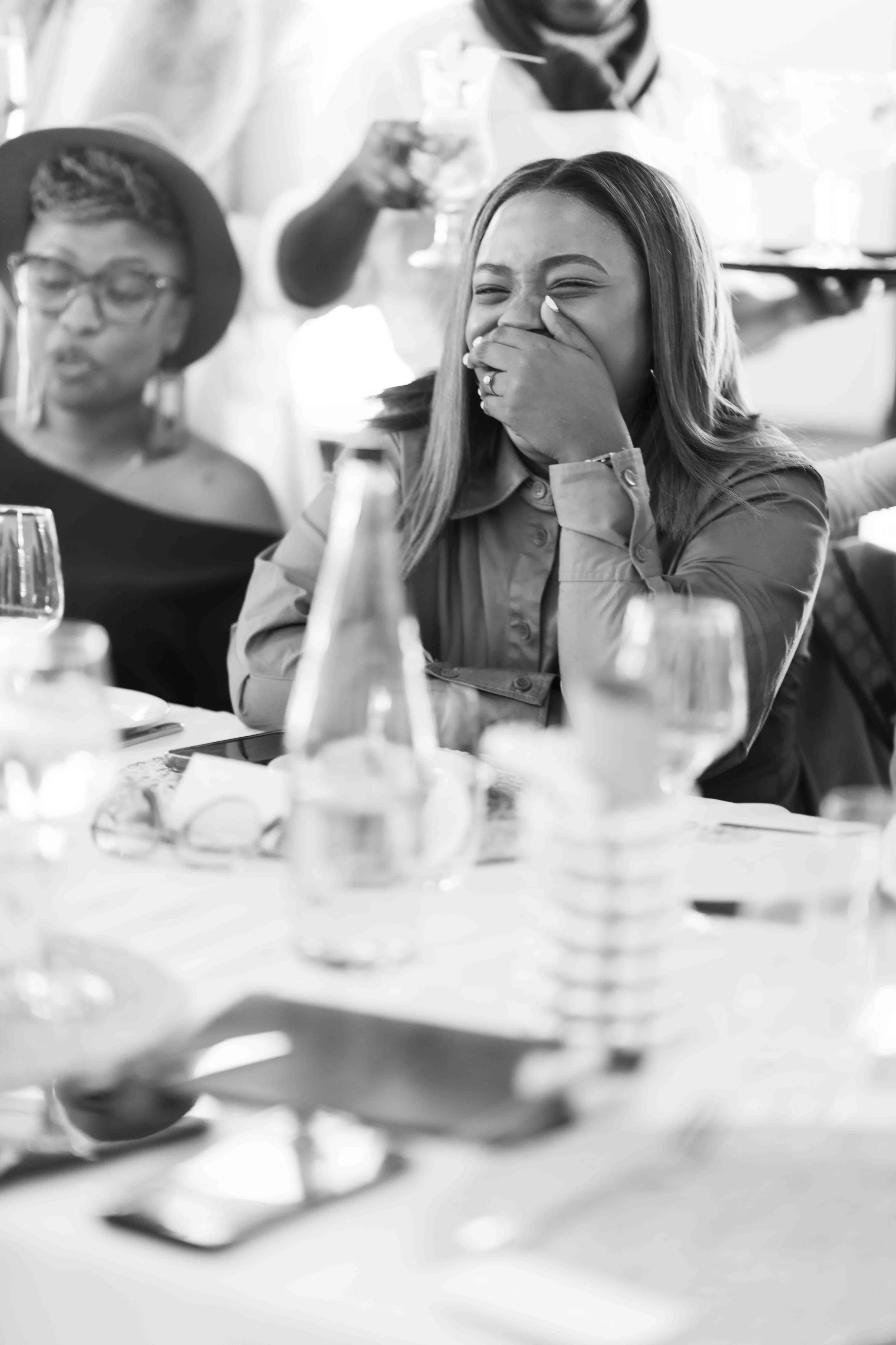 A black and white photograph taken during a baby shower event showing a woman sitting at a table, laughing and covering her mouth with her hand, during a social gathering. Other people and drinks are visible in the background and foreground.
