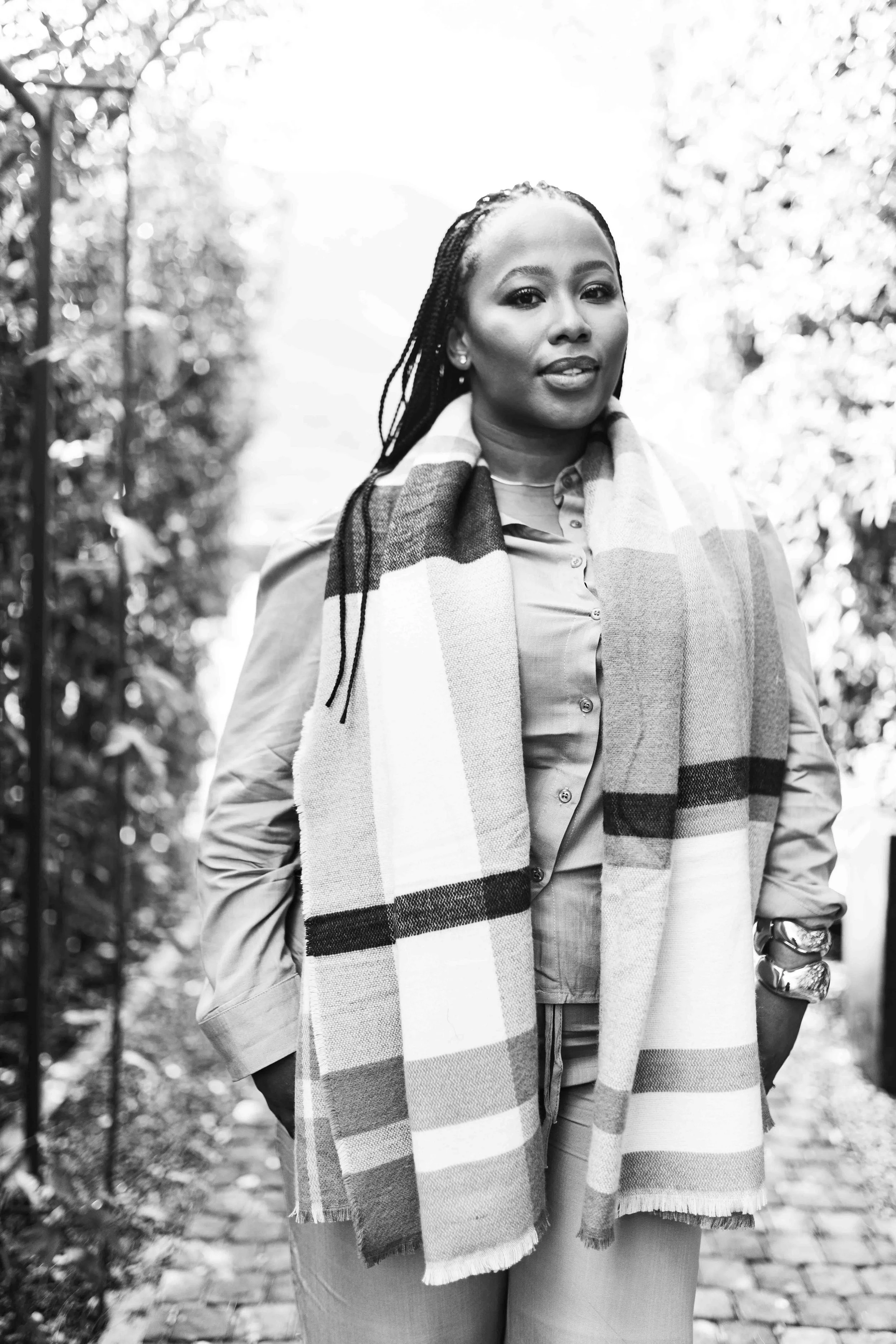 A black and white photograph taken during a baby shower event showing a woman standing outdoors on a cobblestone path, wearing a scarf, a button-up shirt, and a watch.