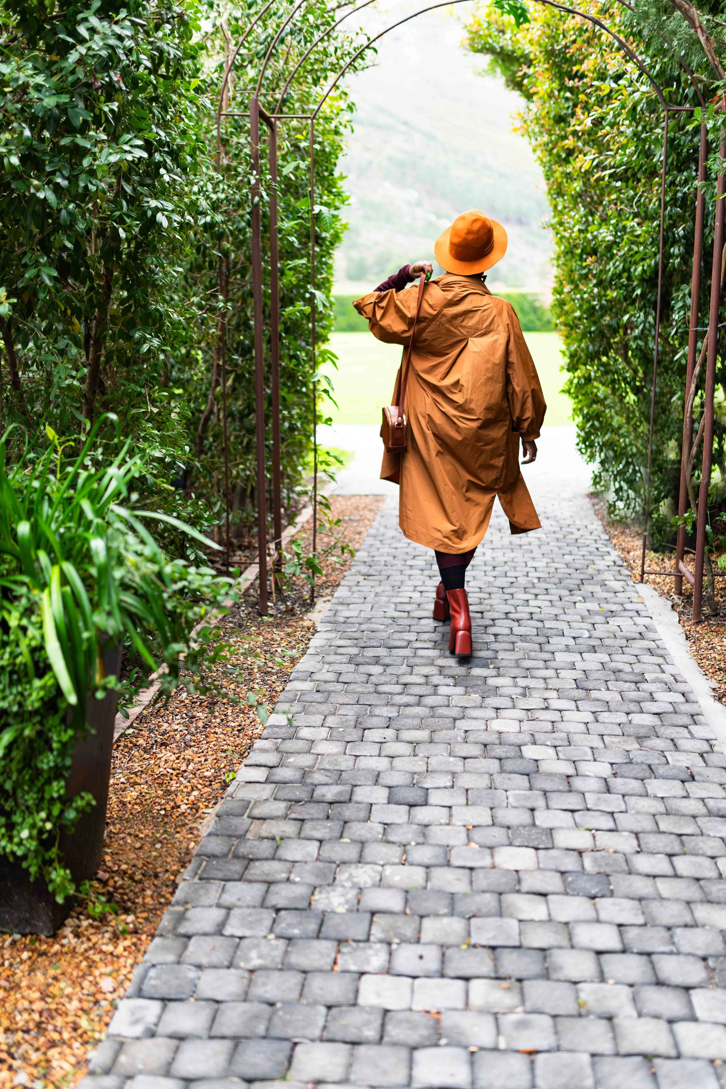 A photograph taken during a baby shower event showing a woman walking through a garden archway on a cobblestone path, wearing a brown coat, orange hat, and red boots.