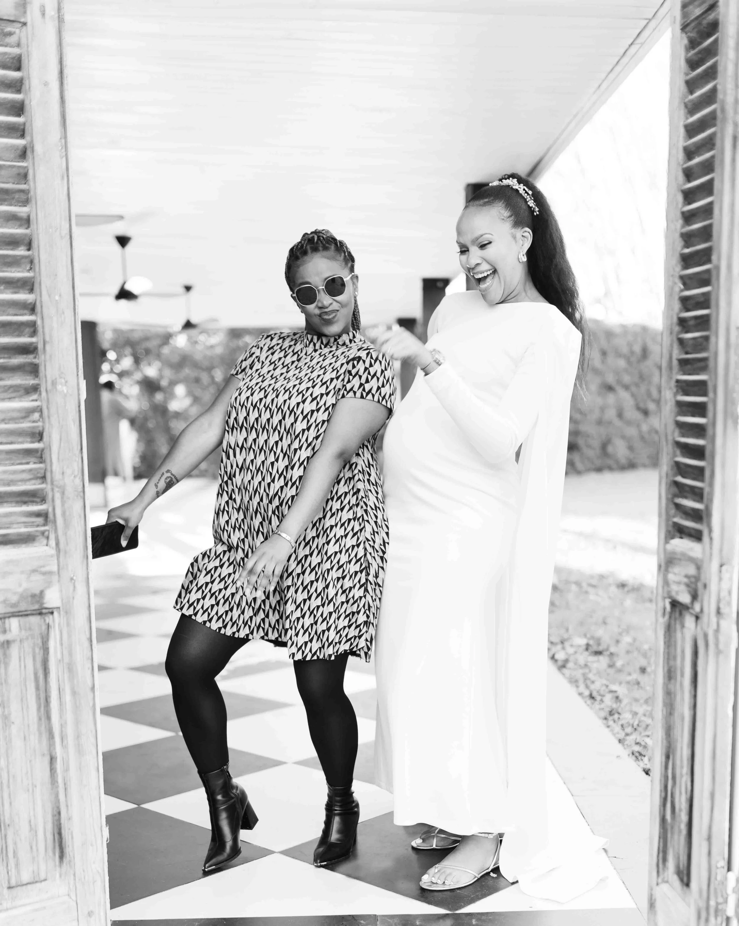 A black and white photograph taken during a baby shower event showing two women smiling and having fun, standing in a doorway with a checkered floor outside.
