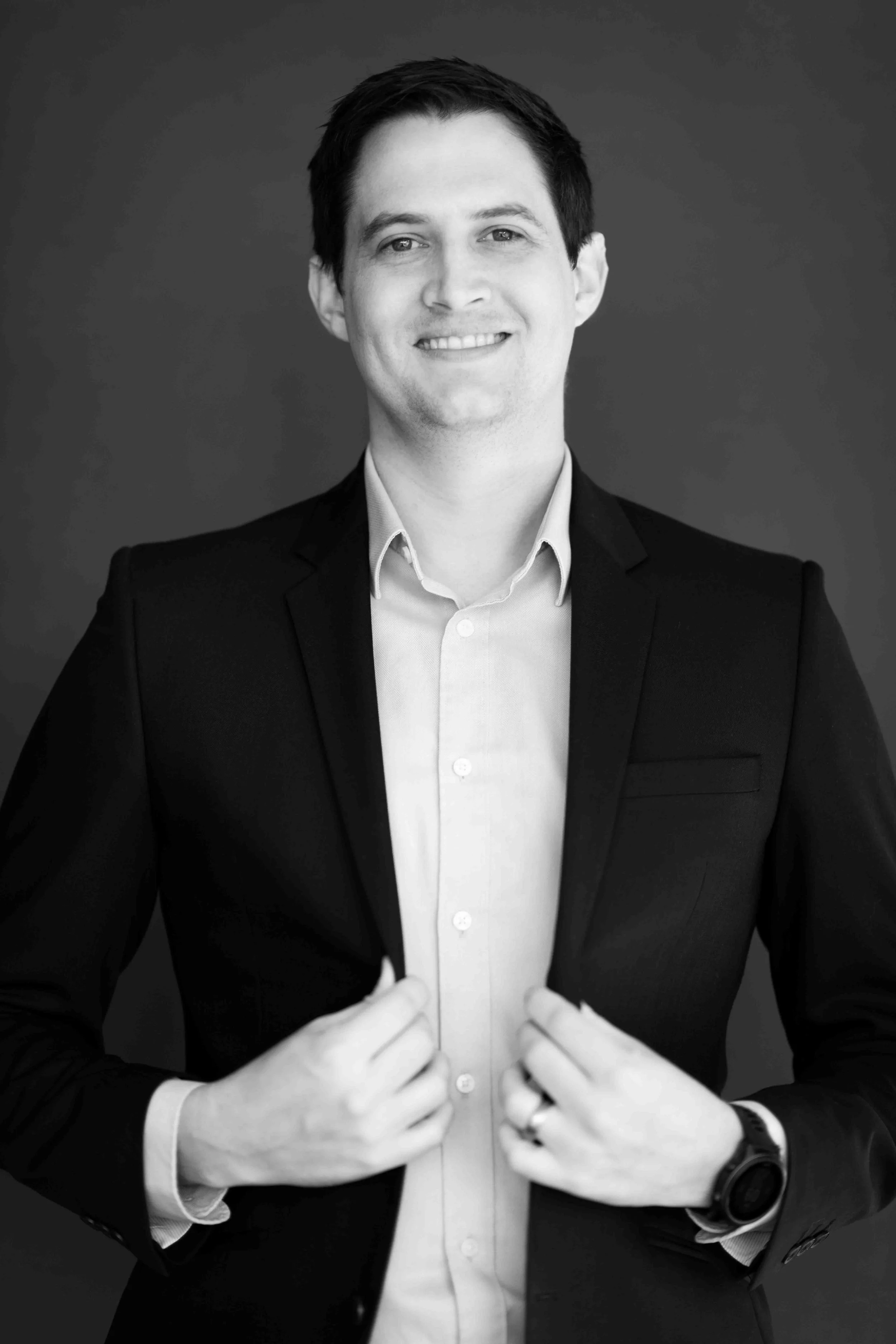 A corporate professional headshot taken in Cape Town in black and white of a smiling man in a suit, holding the lapels of his jacket.