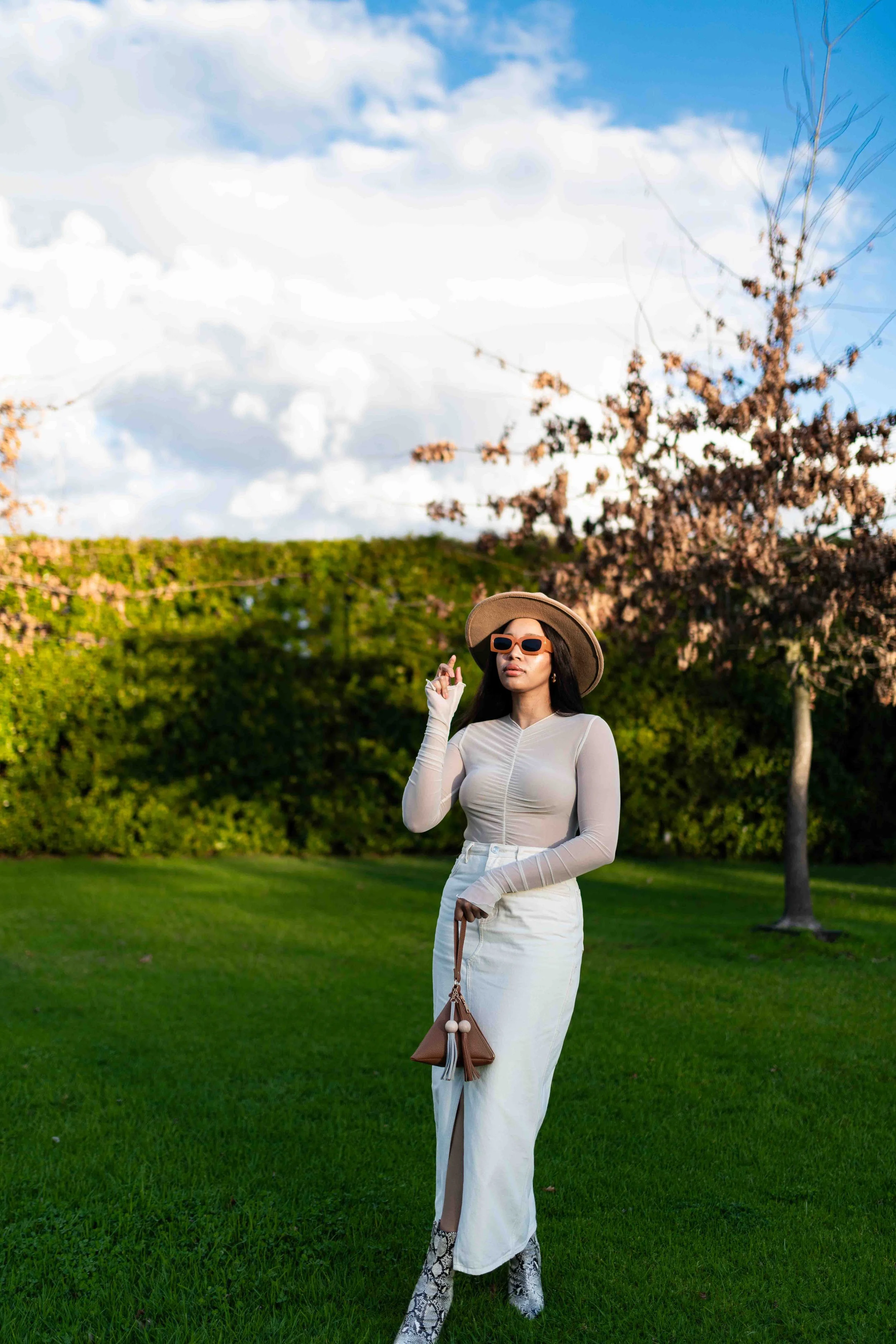 A photograph taken during a baby shower event showing a woman in a beige hat, white sunglasses, and fashionable clothing standing on a grassy field with trees and cloudy sky in the background.