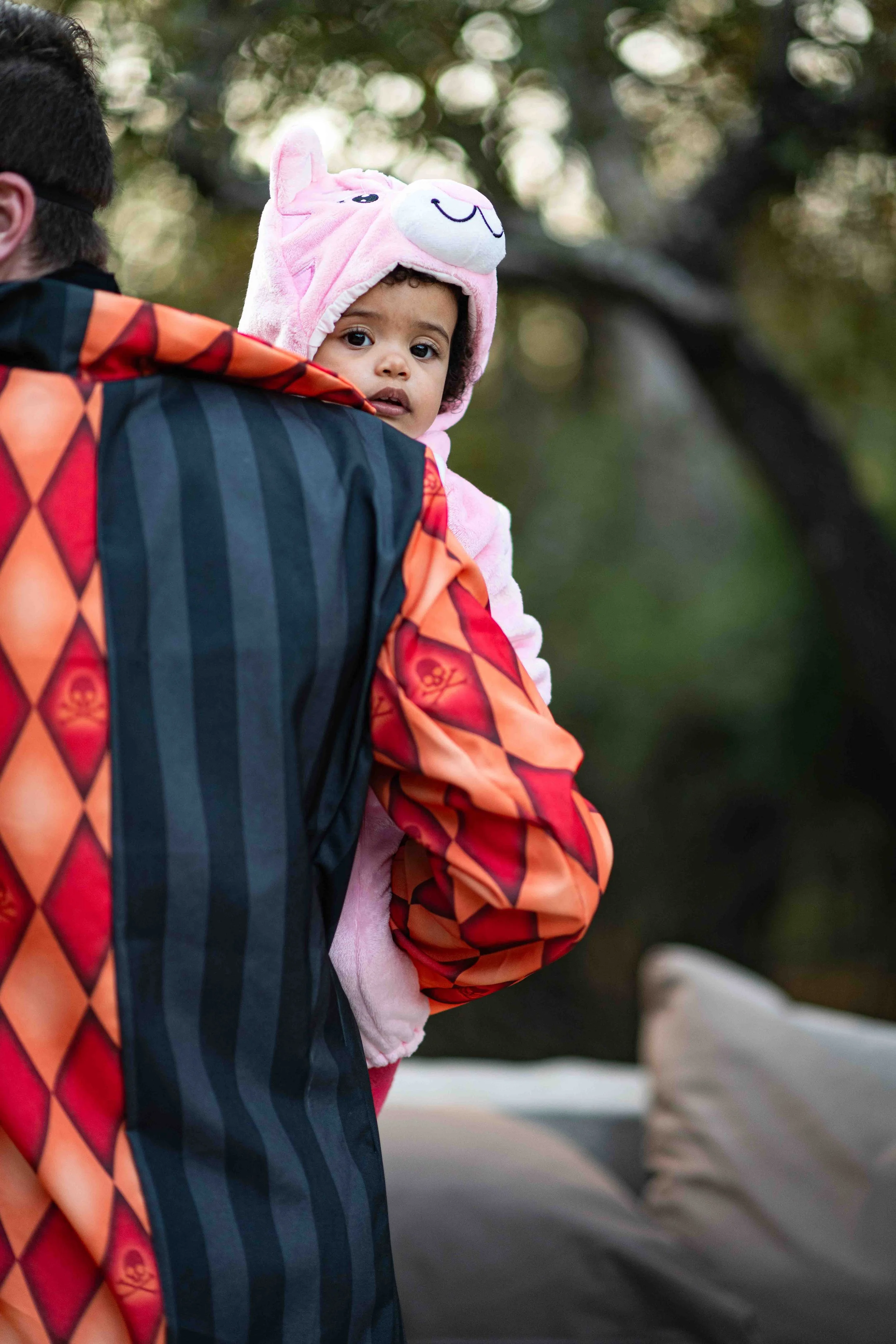A photograph of a young child wearing a pink animal-themed hooded jacket being carried by her father outdoors, with trees in the background at the Kruger National Park