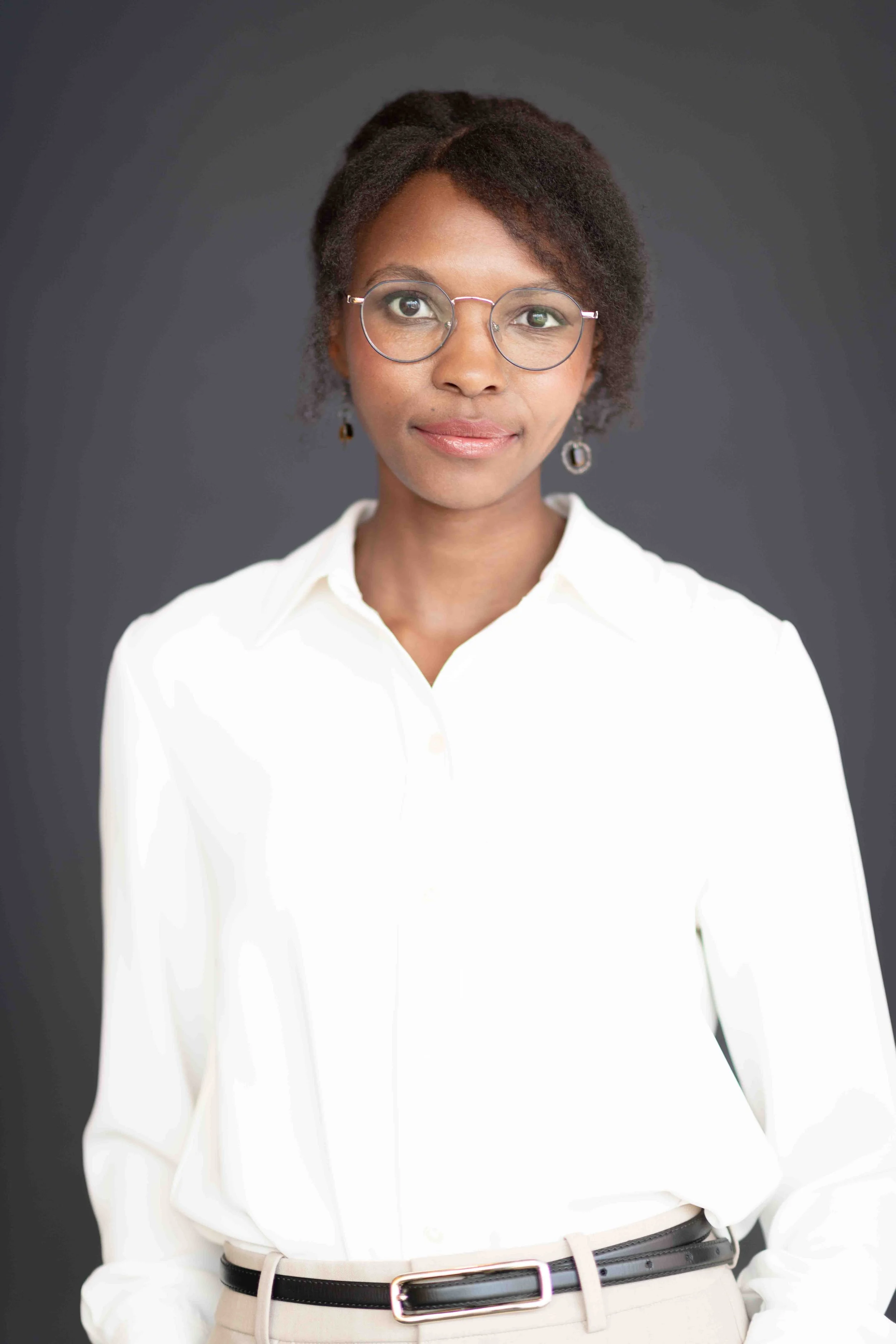 A corporate professional headshot taken in Cape Town of a woman with dark hair, glasses, and earrings, wearing a white blouse and beige pants, standing against a dark grey background.