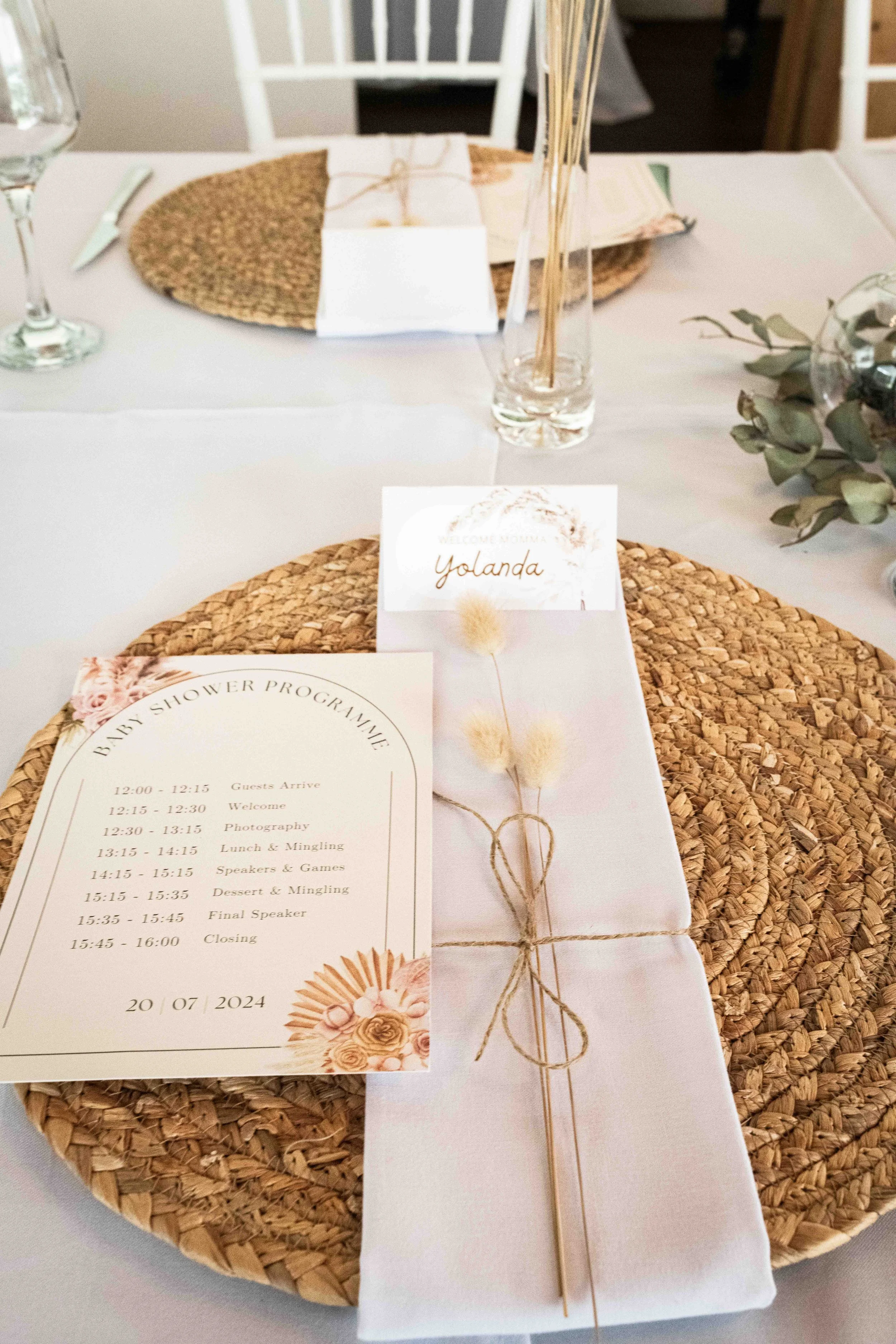 A photograph taken during a baby shower event showing an elegant table setting, with a woven placemat, a welcome card, a shower program, decorated with dried flowers and natural accents.