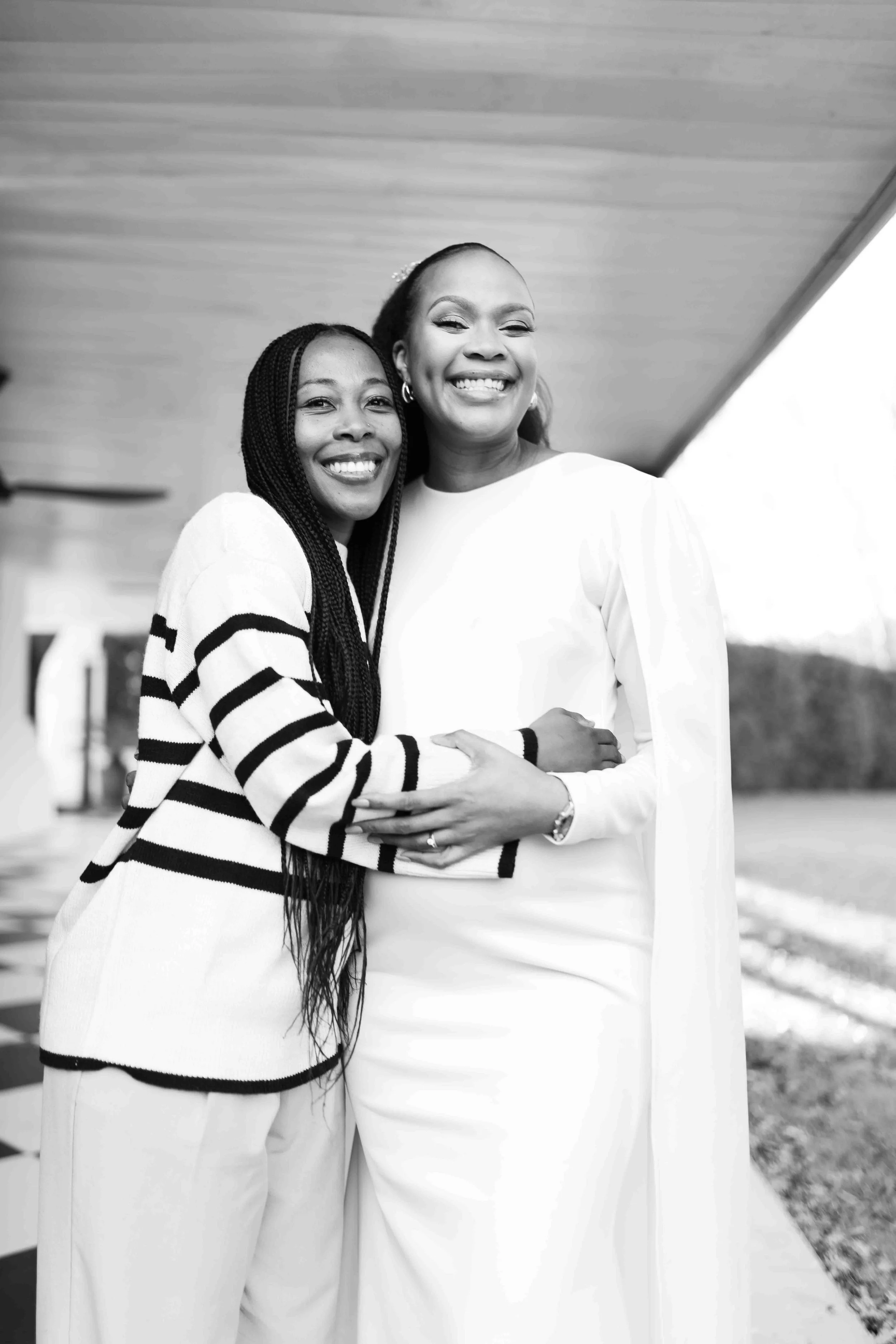 A black and white photograph taken during a baby shower event showing two women smiling and embracing, outdoors.