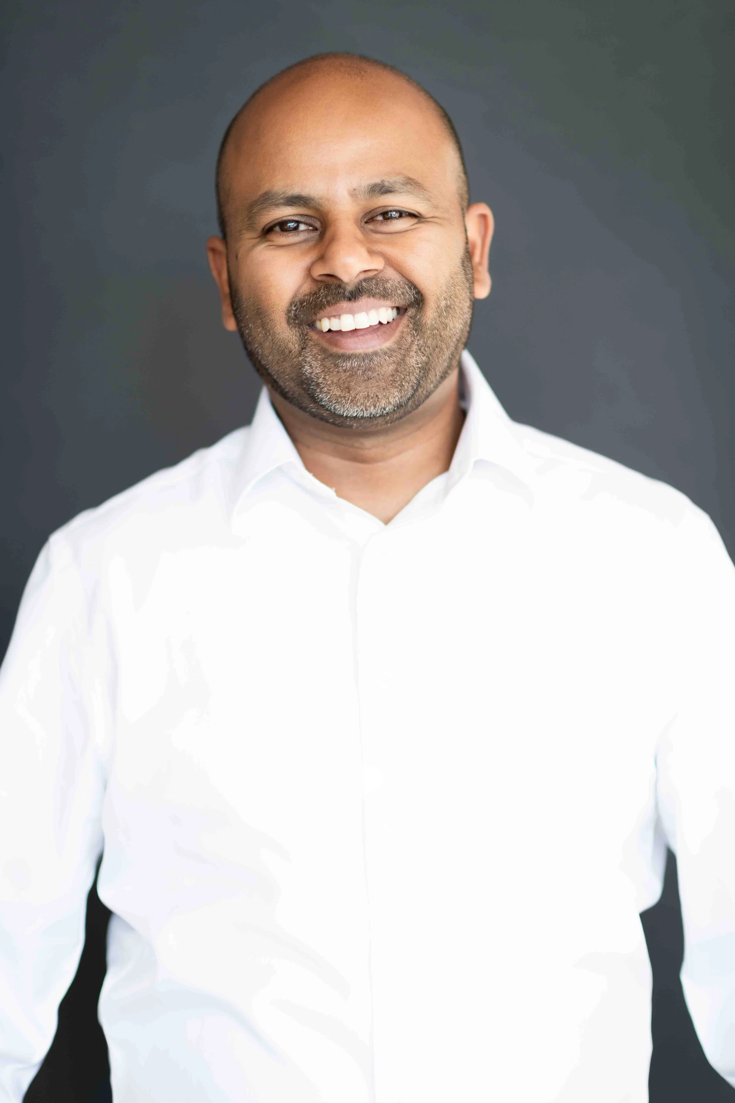 A corporate professional headshot taken in Cape Town of a smiling man with a beard and bald head, wearing a white dress shirt, standing against a dark background.