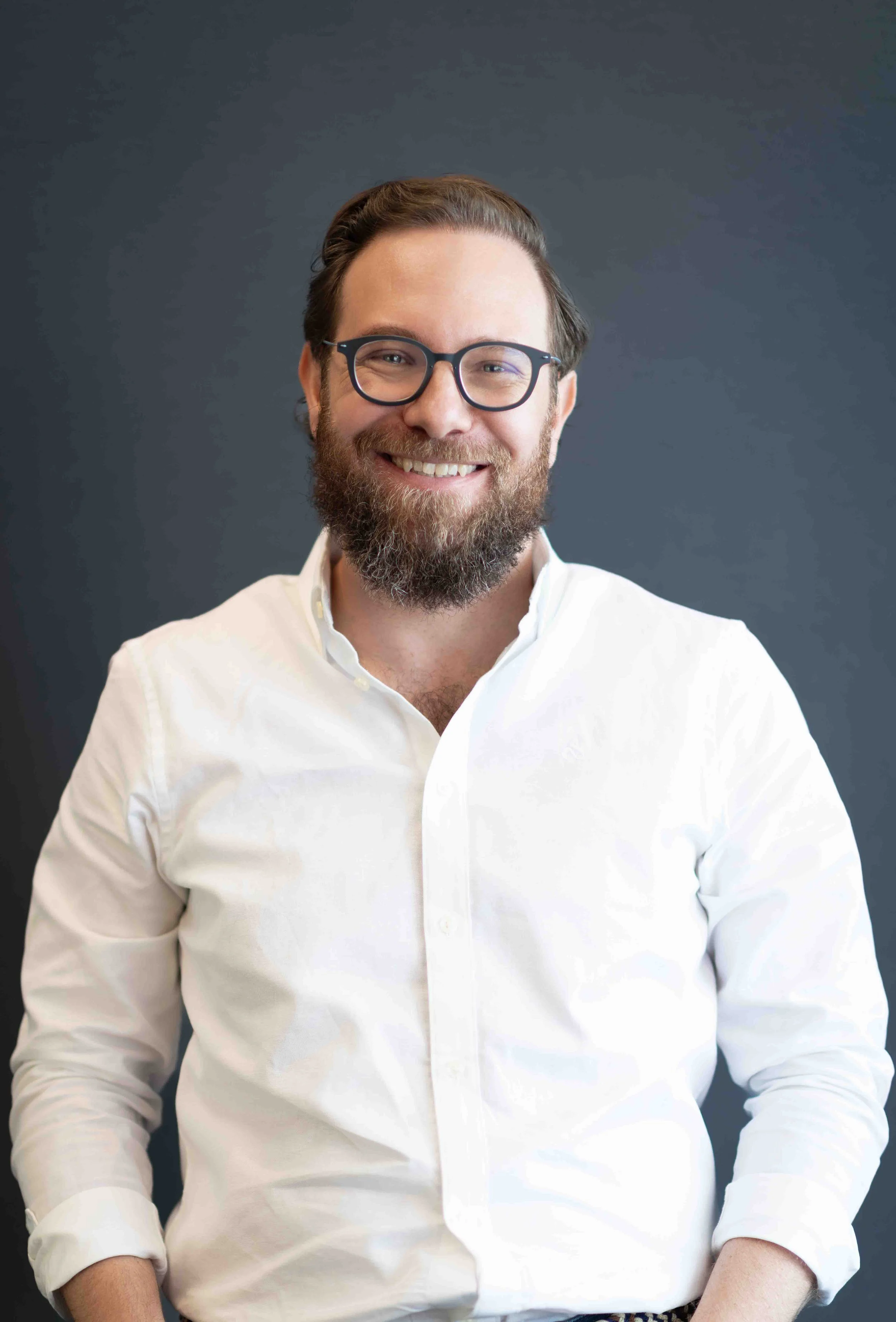 A corporate professional headshot taken in Johannesburg of a man with glasses and a beard smiling in front of a dark background, wearing a white button-up shirt.