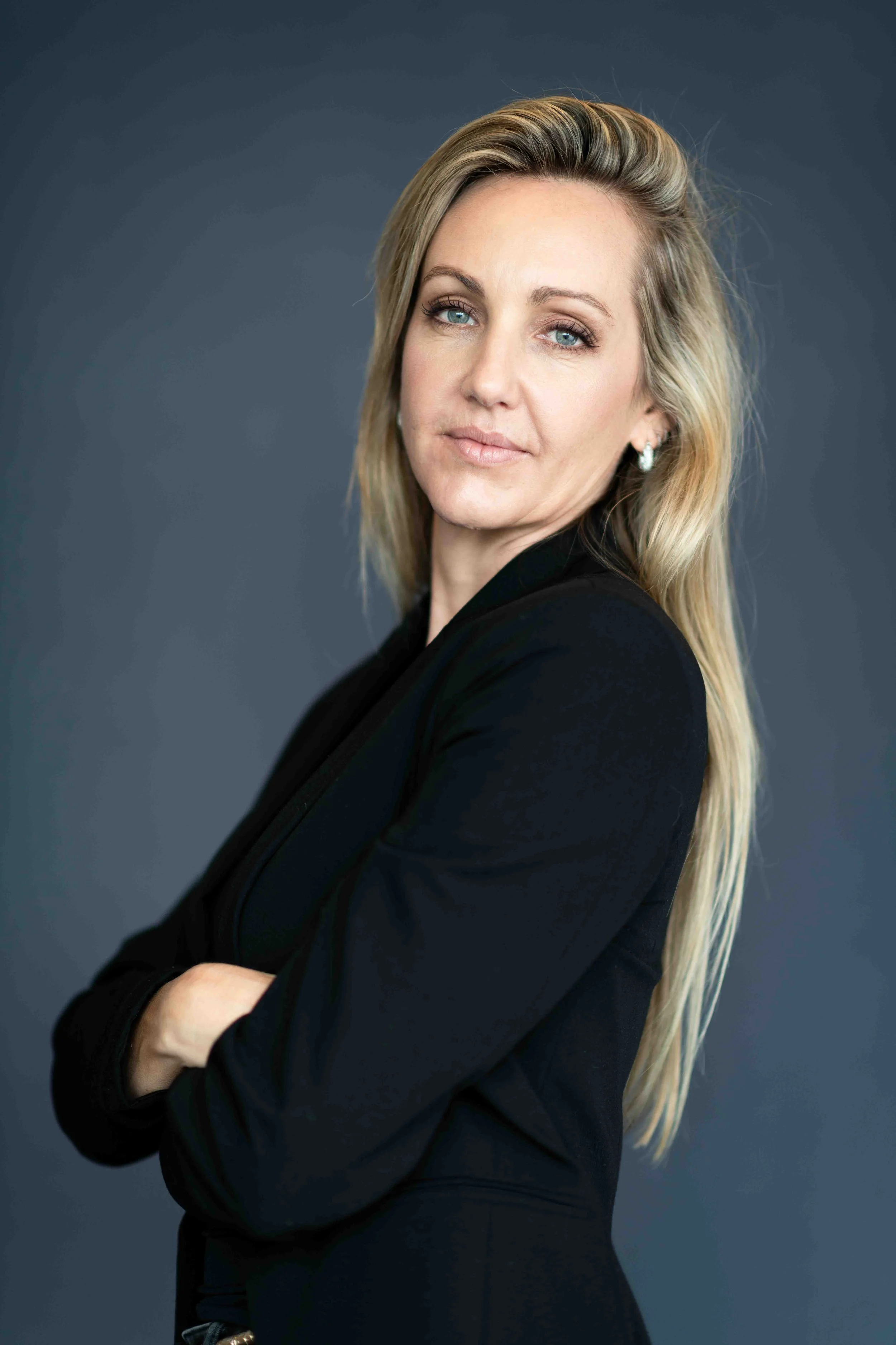 A corporate professional headshot taken in Cape Town of a woman with long blonde hair, wearing a black blazer, standing against a dark grey background with arms crossed and a confident expression.