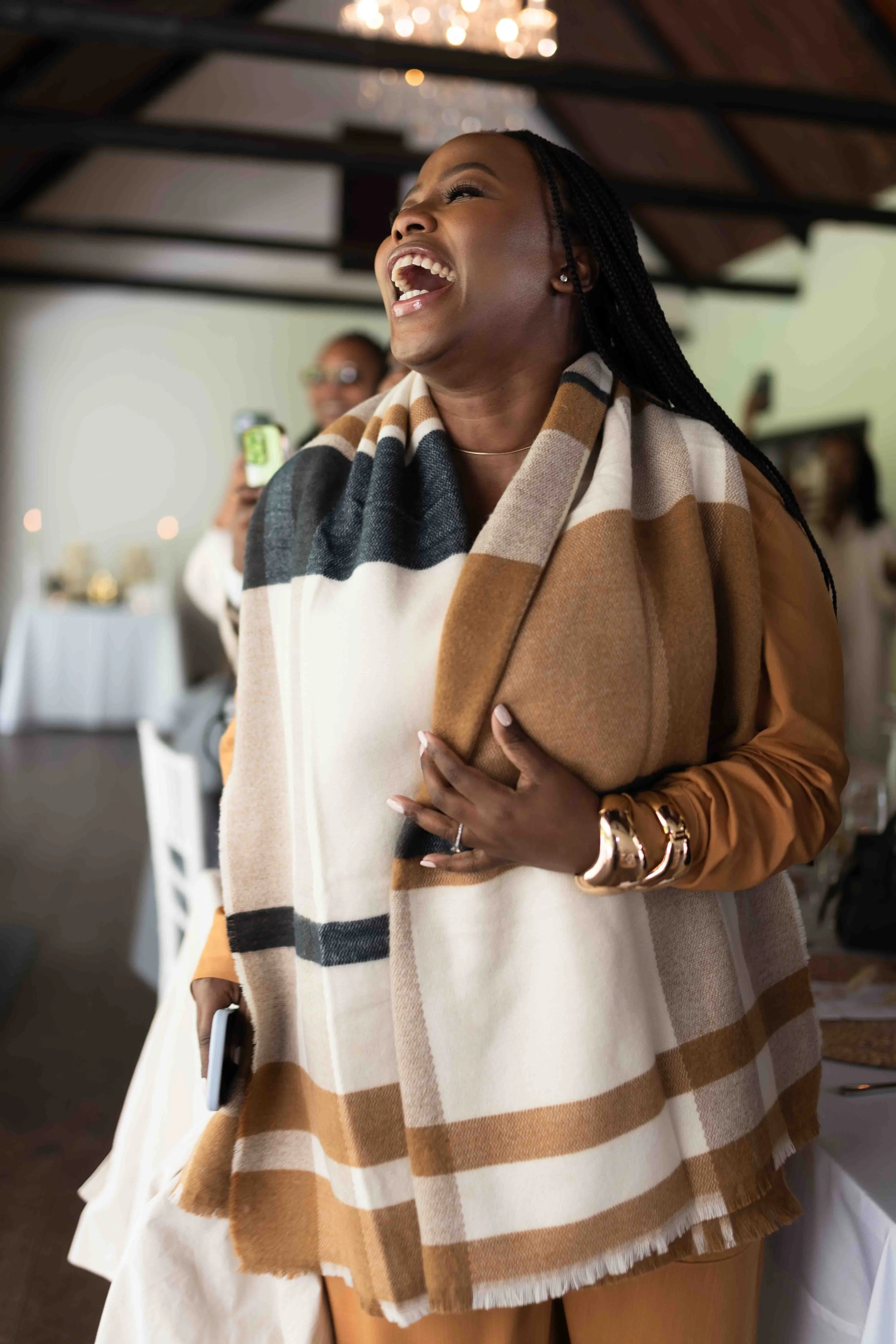 A photograph taken during a baby shower event showing a happy woman laughing, wearing a beige and black plaid scarf and gold jewellery, standing indoors at a social gathering.