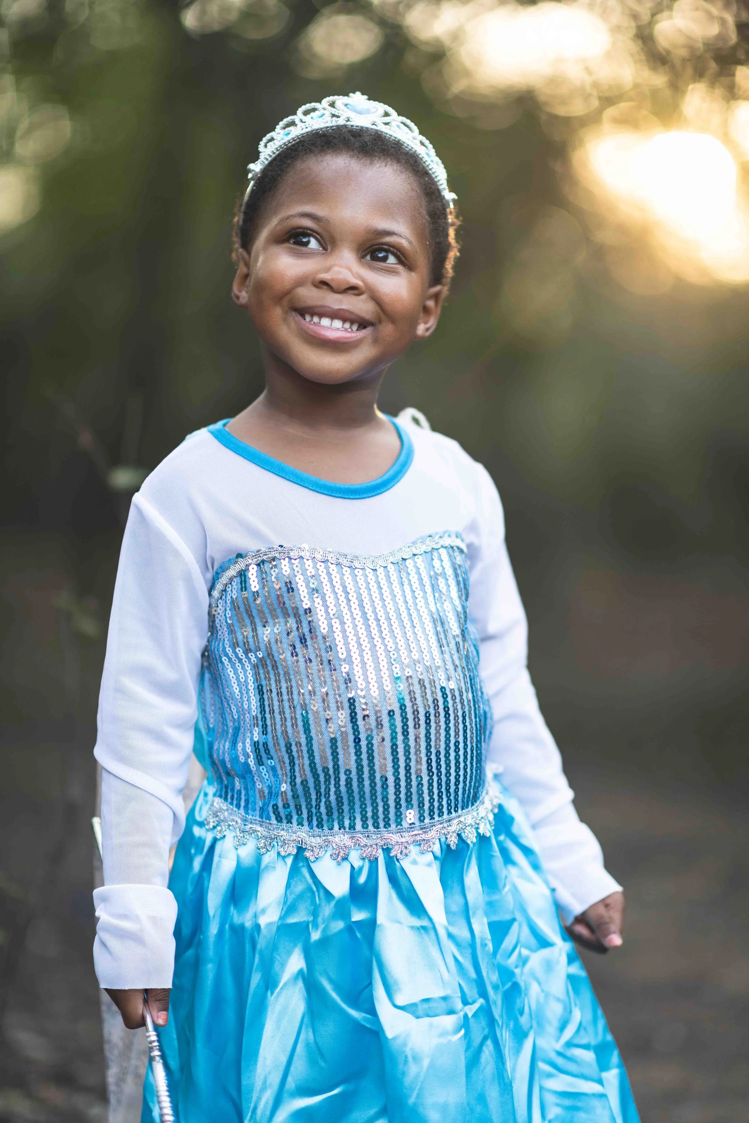 A portrait photograph of a young girl in costumes at a birthday party outdoors at the Kruger National Park, wearing a blue and white princess dress and tiara, smiling outdoors at sunset.