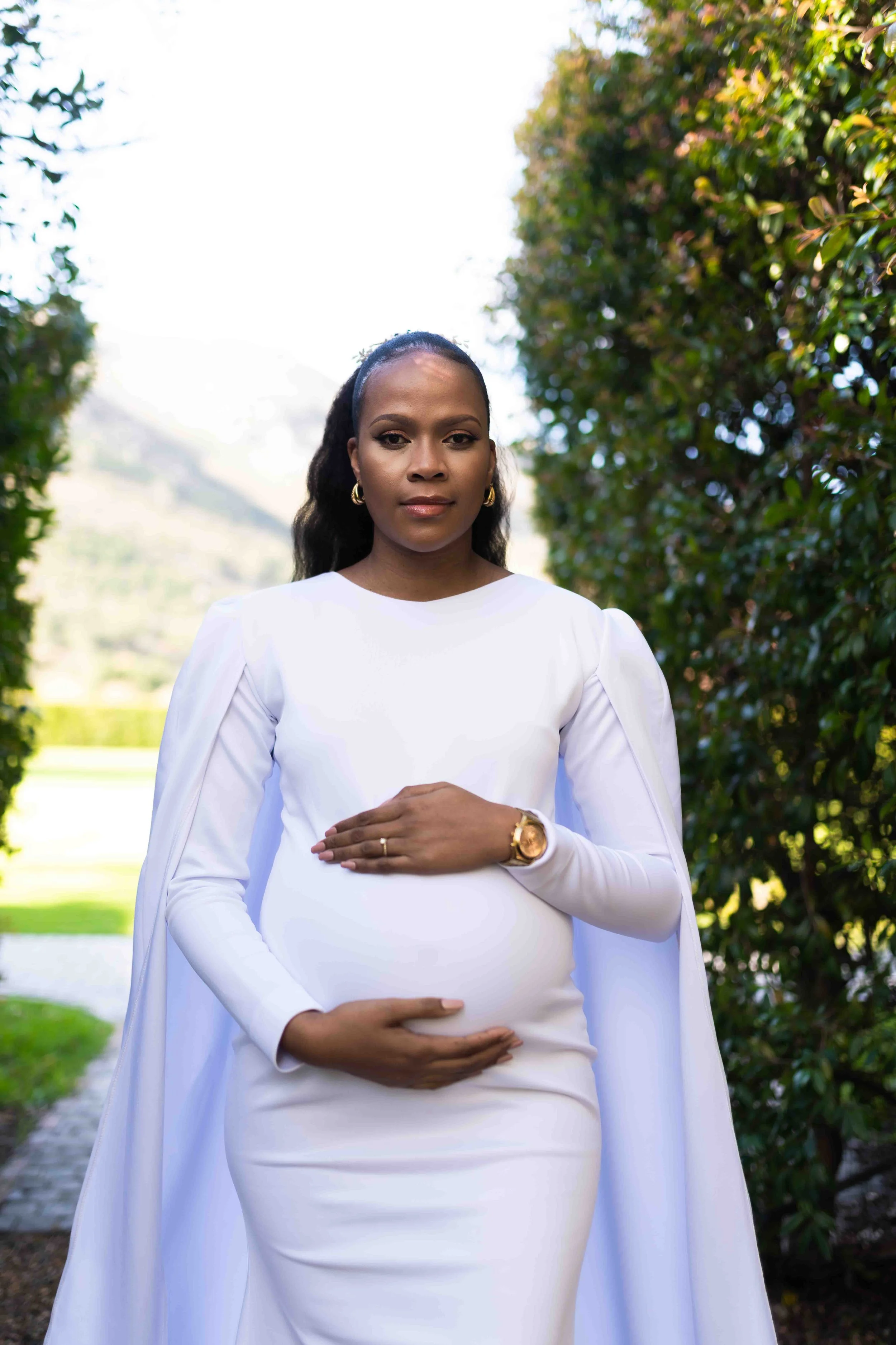 A photograph taken during a baby shower event showing a the guest of honour, dressed in a white gown with a cape, holding her pregnant belly, standing outdoors between greenery, with mountains in the background, looking at the camera.