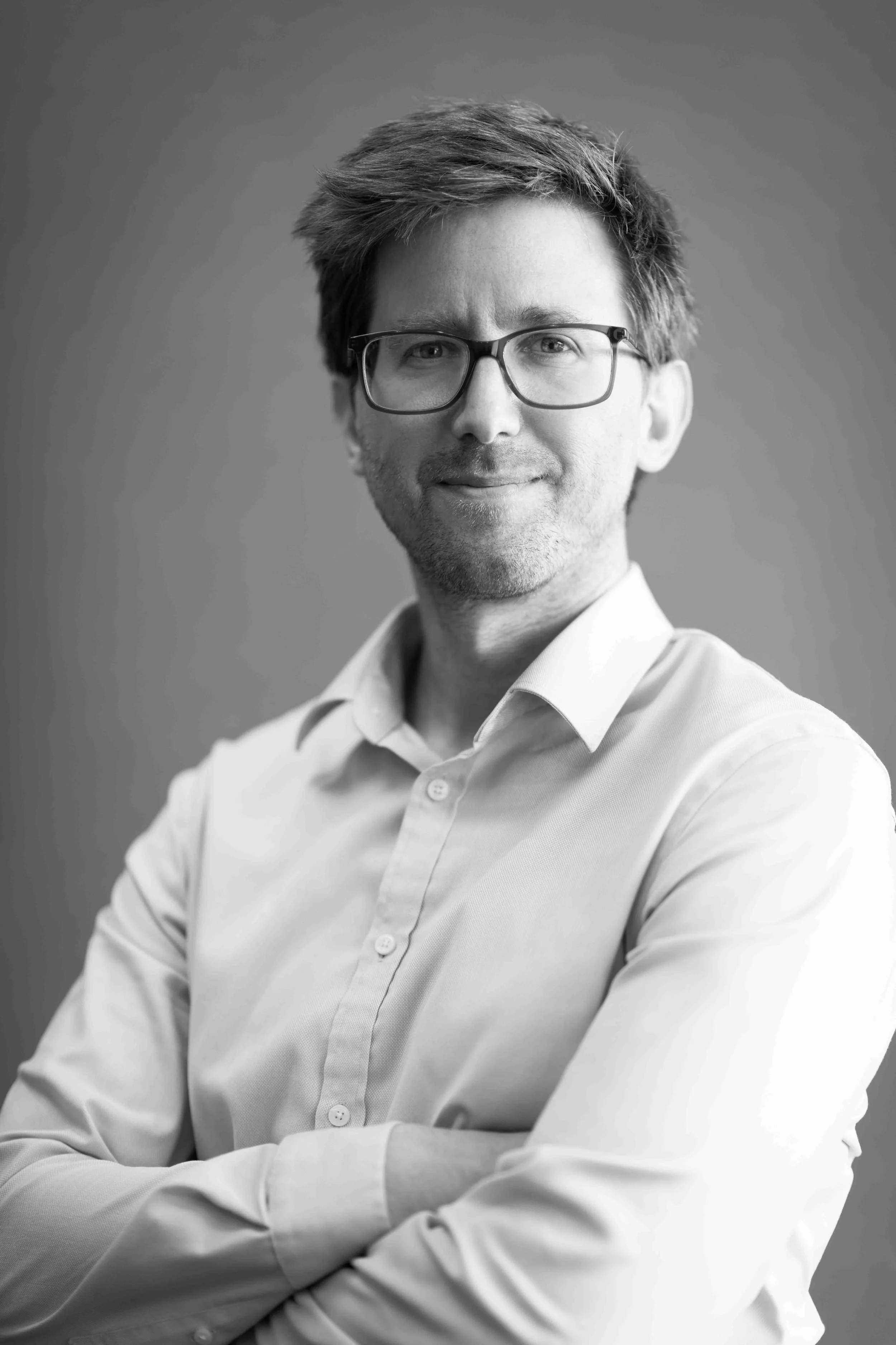 A corporate professional headshot taken in Johannesburg in black and white of a man with glasses, smiling, wearing a light-coloured collared shirt, facing slightly to the right with arms crossed.