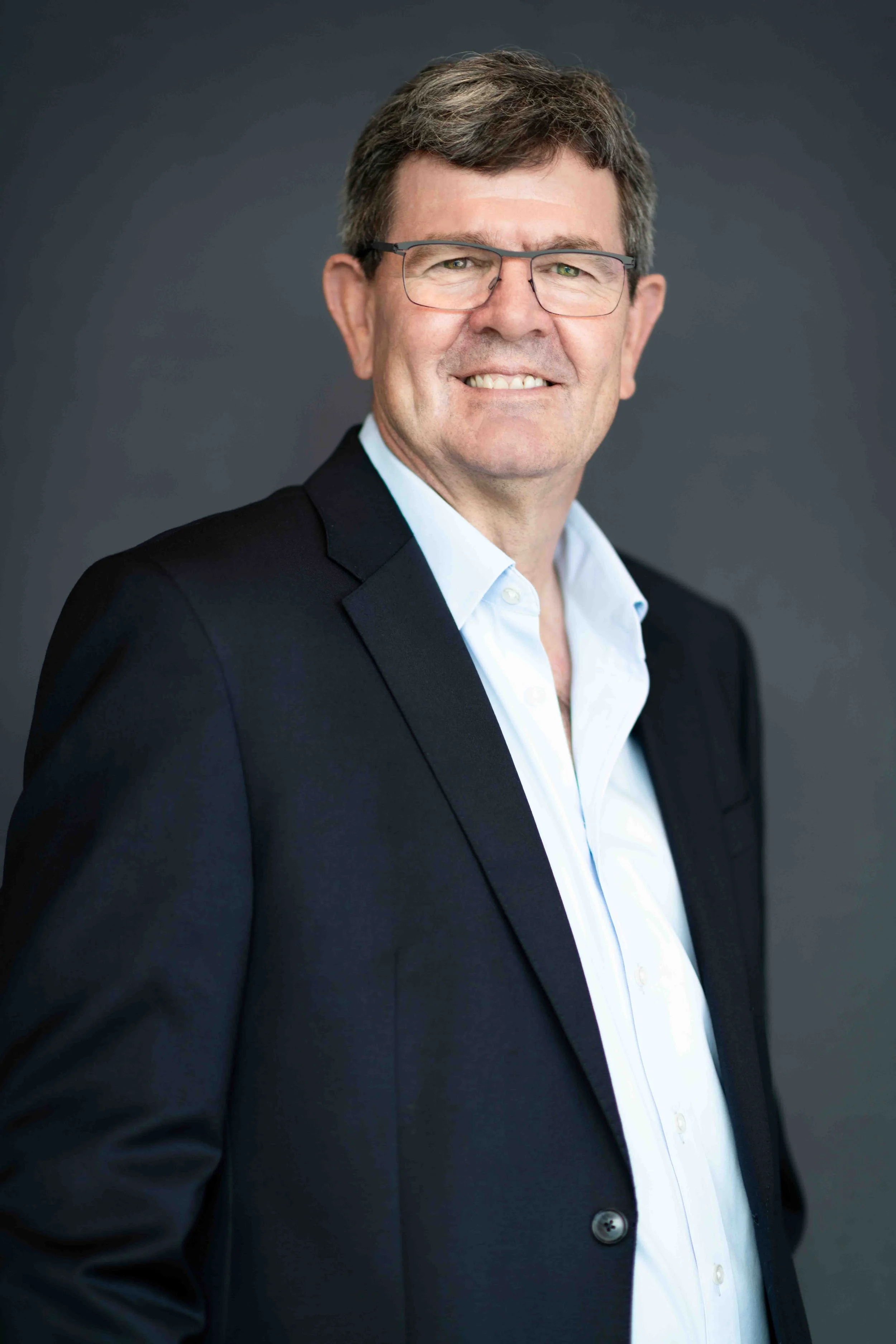 A corporate professional headshot taken in Cape Town of a man with glasses, wearing a black blazer and white shirt, smiling in front of a dark grey background.