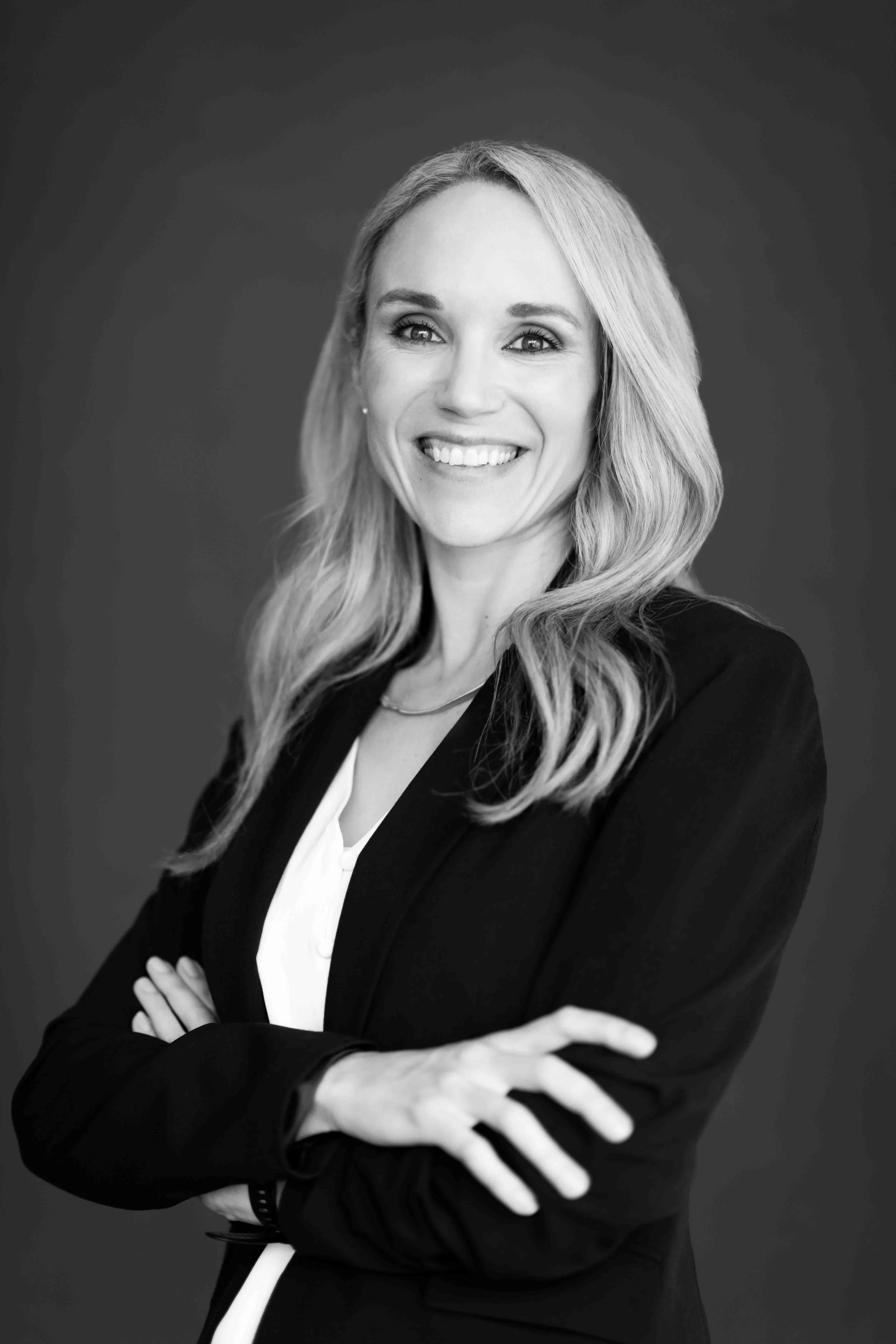 A corporate professional headshot taken in Cape Town in black and white of a woman with long wavy hair, smiling, wearing a dark blazer and a white blouse, arms crossed.
