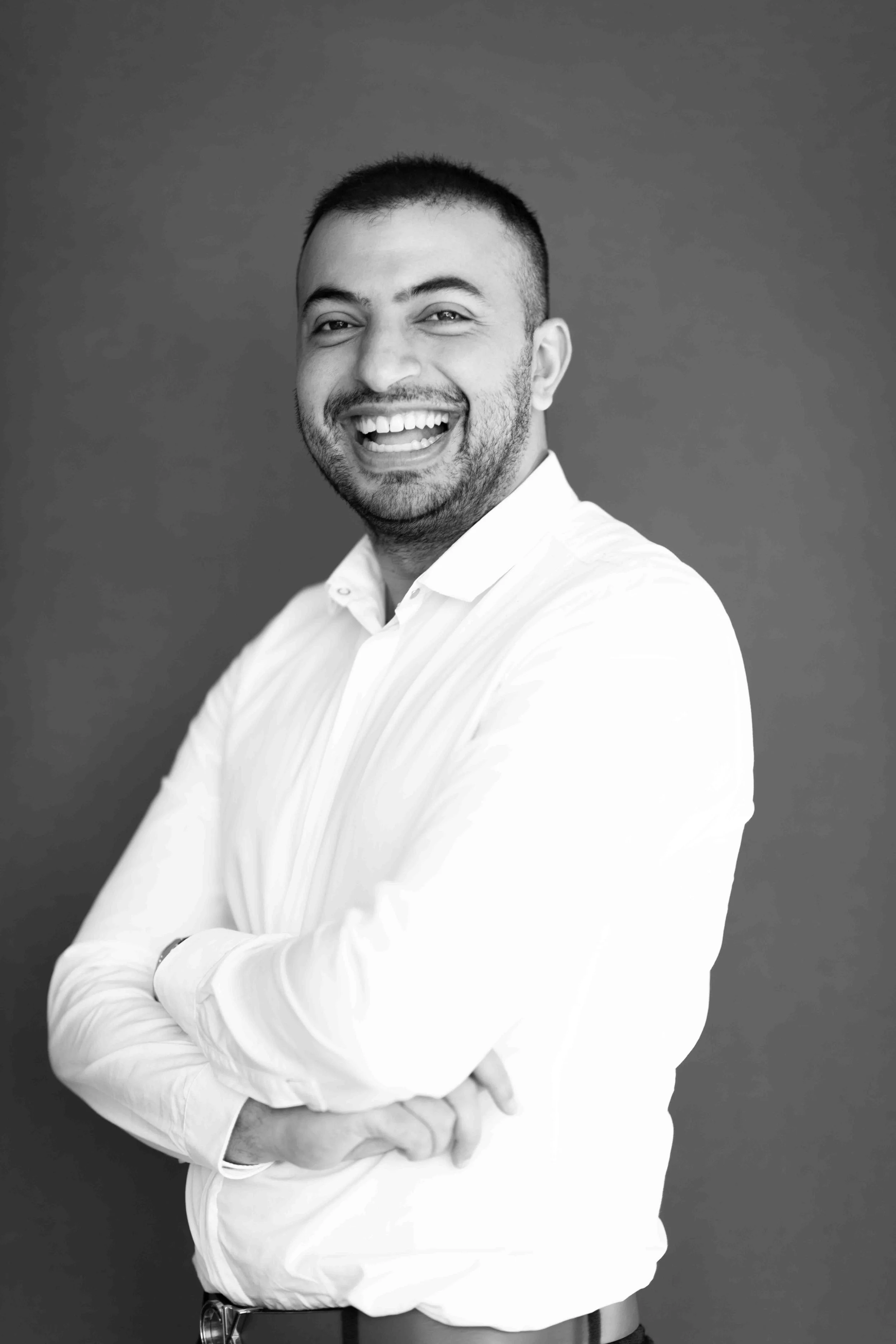 A corporate professional headshot taken in Cape Town in black and white of a smiling man with short dark hair and beard, wearing a white shirt, standing with arms crossed against a plain background.