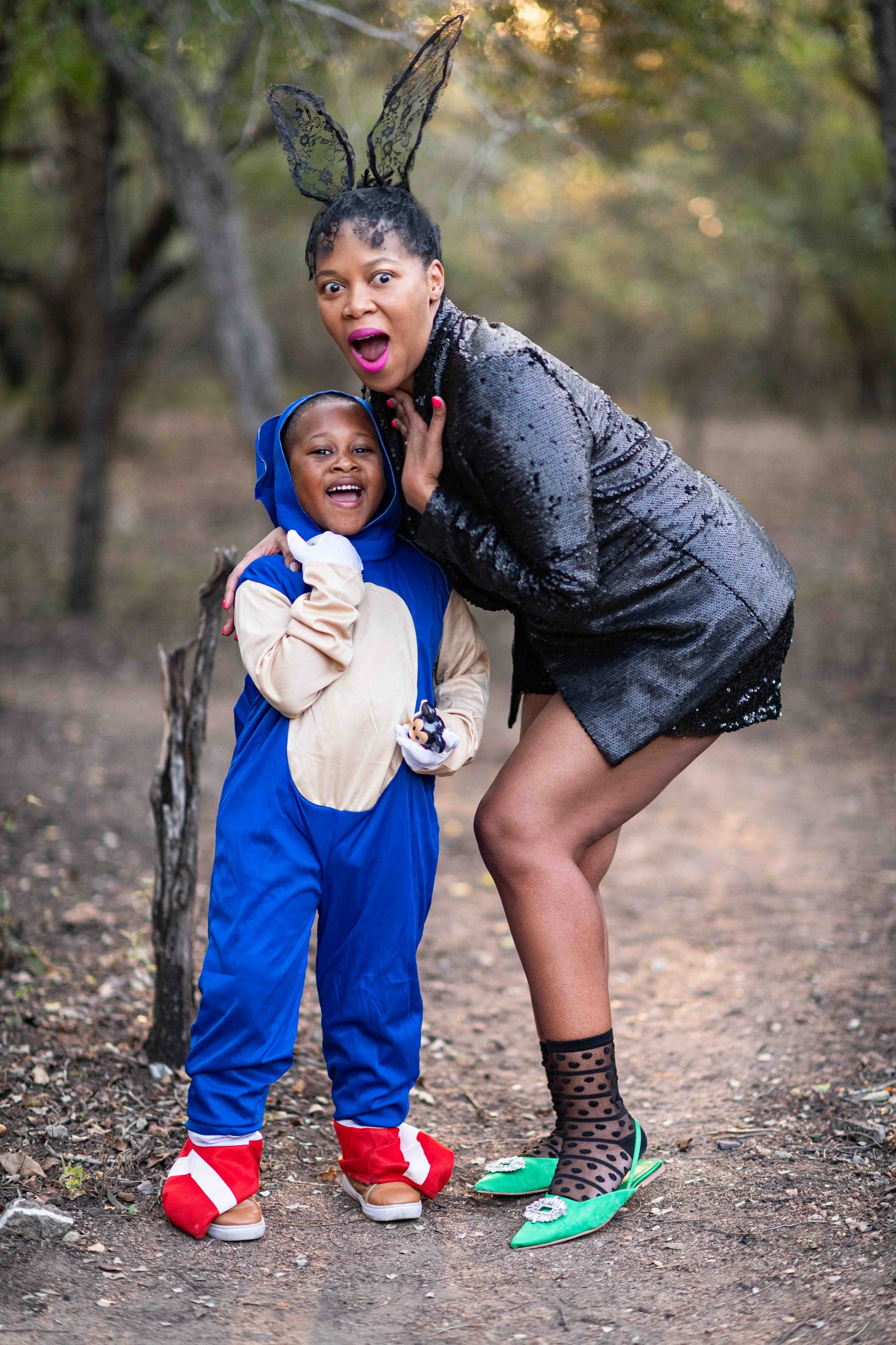 A photoshoot of a mother and son in costumes at a birthday party outdoors at the Kruger National Park. The woman in a black sequin dress and bunny ears, and the child in a blue and beige animal onesie, both smiling and posing for the camera.