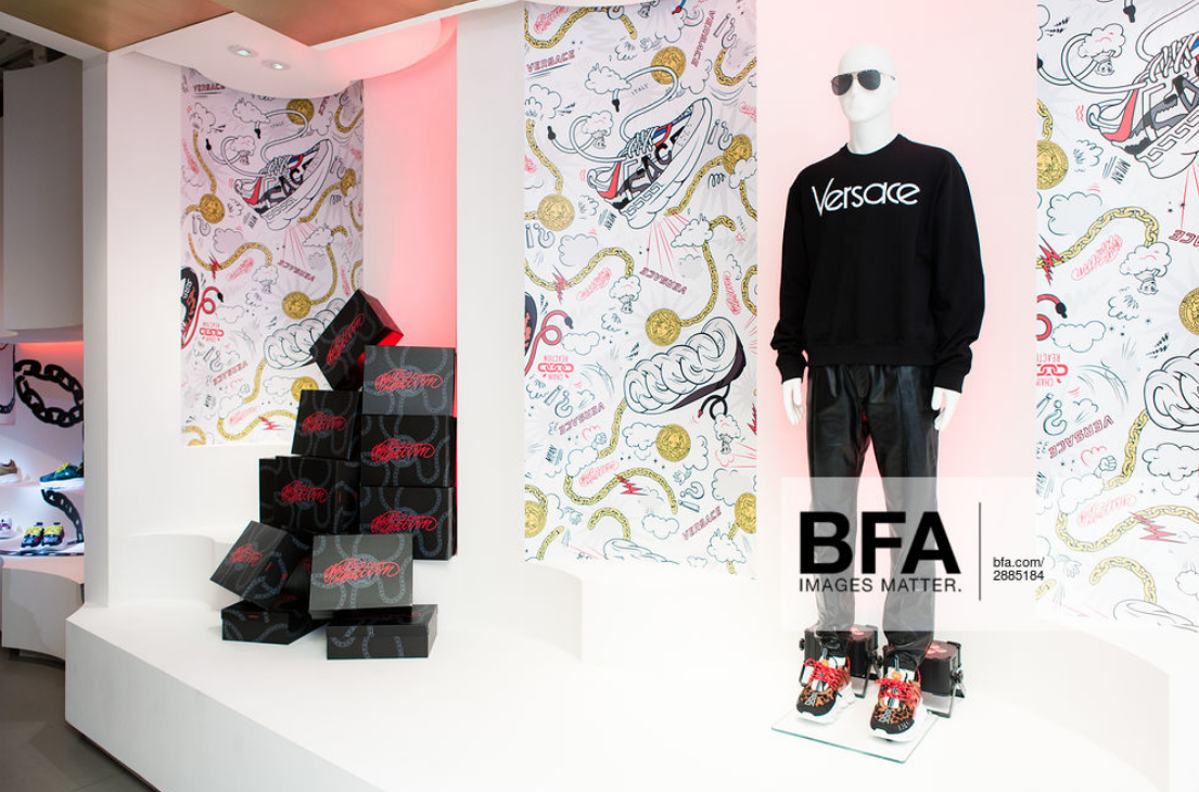 Fashion display with a mannequin wearing a black Versace sweatshirt, gray pants, and colorful sneakers, set in a store with patterned wallpaper and a stack of black boxes with red text.