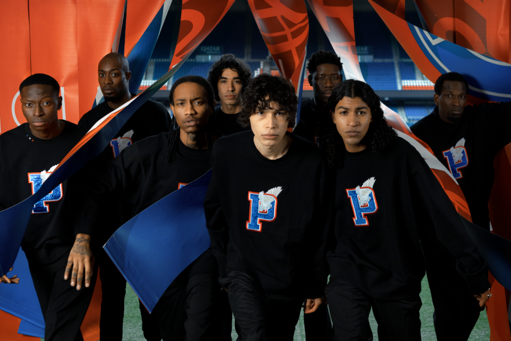 Group of seven young athletes at a sports stadium, wearing black sweatshirts with a stylized letter 'P' and a feather, posing with a large blue and orange banner, 3PARADIS, PSG, Graphic design
