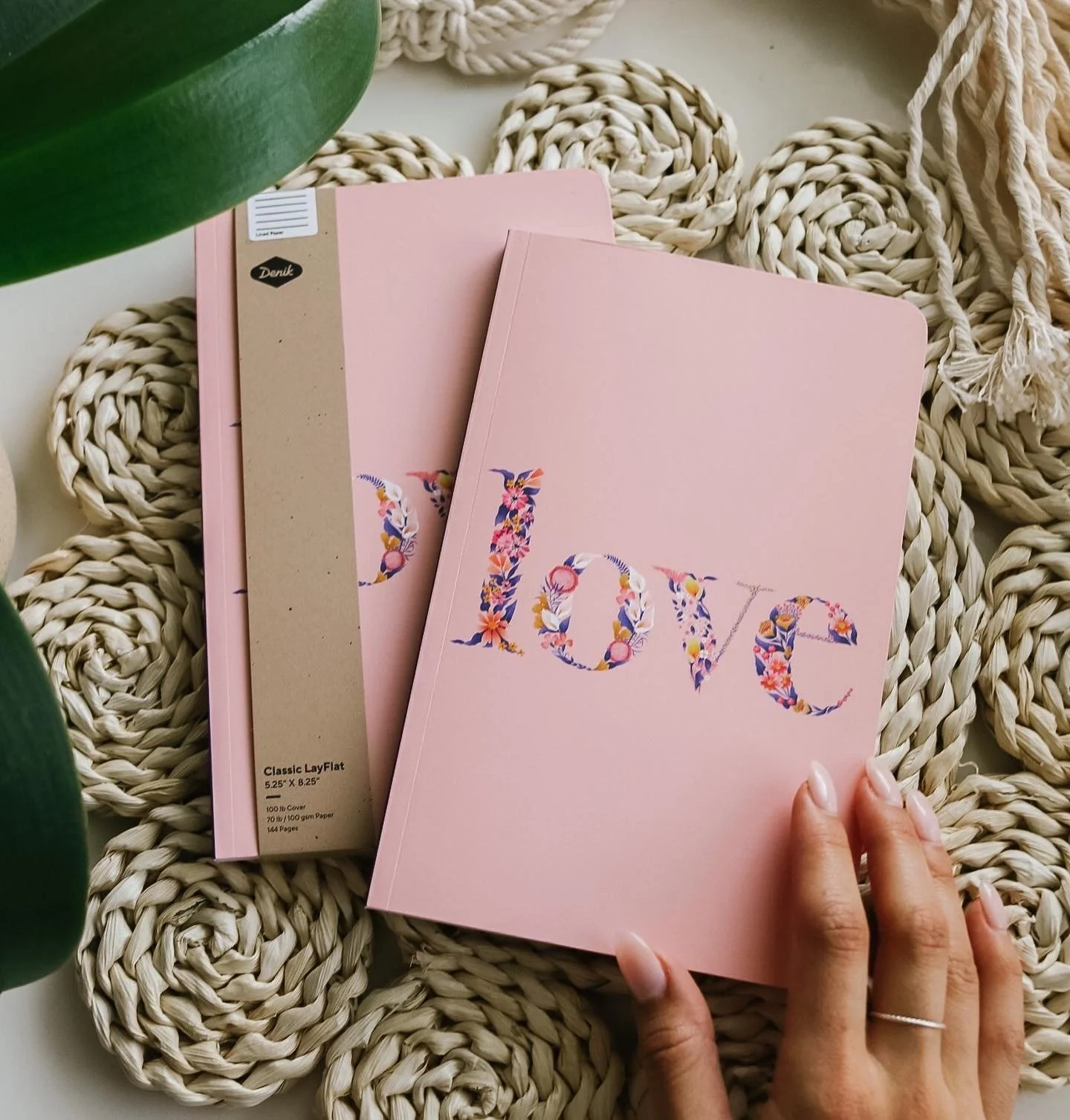 Pink notebook with the word "love" written in floral letters, surrounded by woven placemats and a partial hand with a ring.