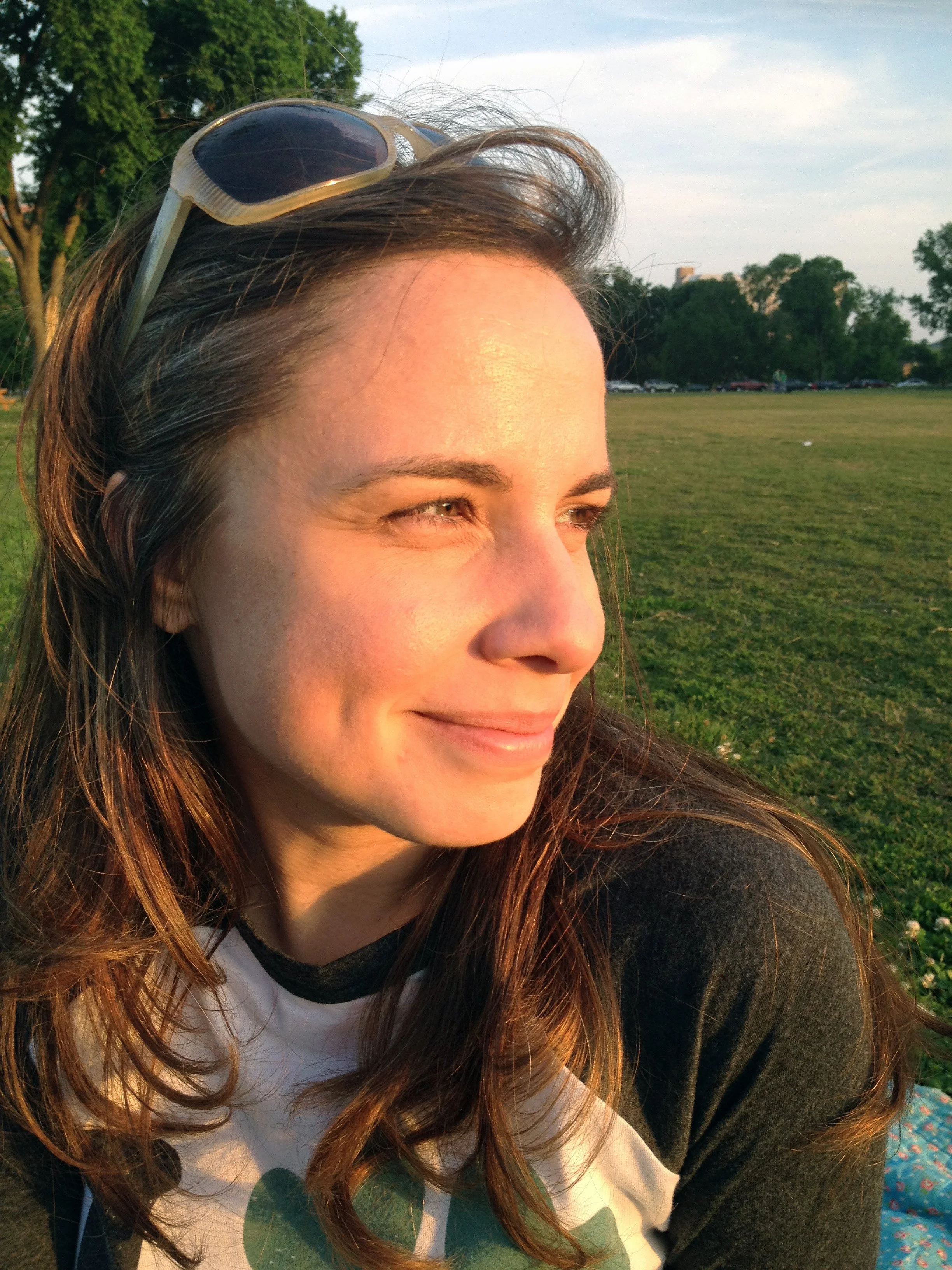 Close-up of a woman with long brown hair, wearing sunglasses on her head, smiling and looking to the side outdoors with a green field and trees in the background during daylight.