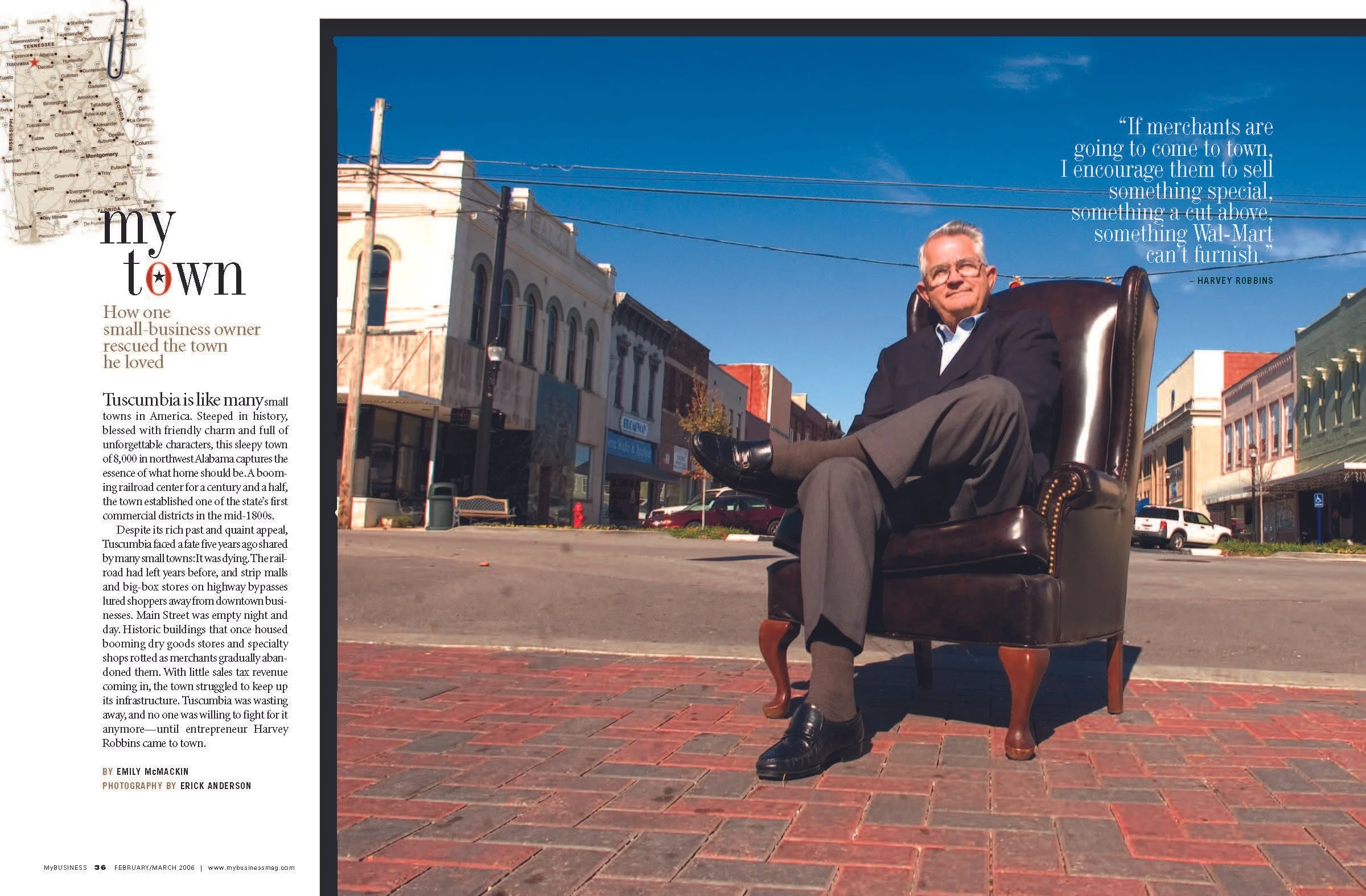 A man with gray hair wearing glasses, a dark blazer, and gray pants, sitting in a leather armchair outdoors on a brick sidewalk in a small town. The town has historic buildings, utility poles, and parked cars; the sky is clear and blue.