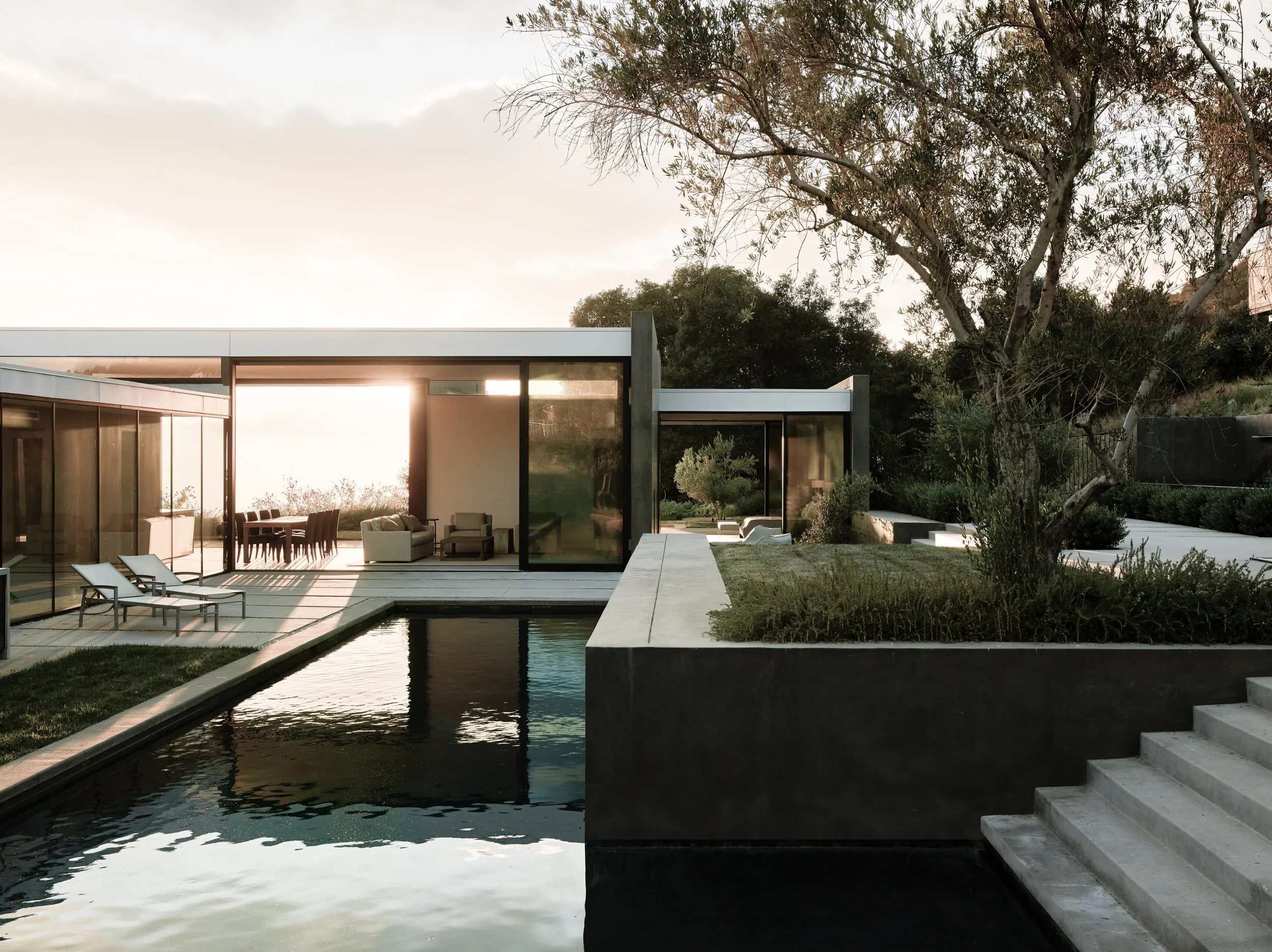 The setting sun shining through an open-air, modern residential home and glittering swimming pool, designed by Walker Workshop in Los Angeles, California.