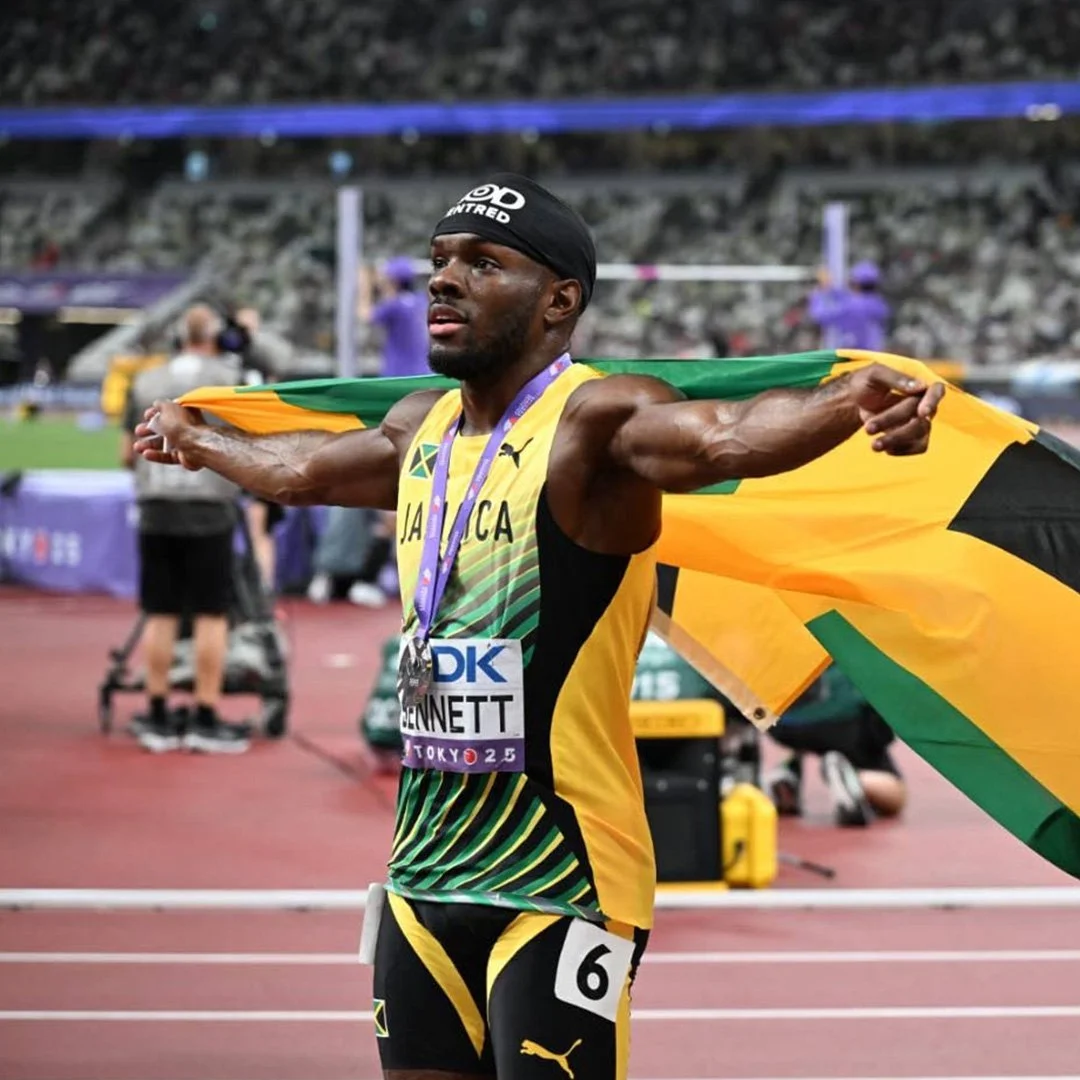A male athlete celebrating with a Jamaican flag draped around his shoulders after winning a race at a track and field event. He is wearing a yellow, black, and green uniform with the number 6 and a medal around his neck.