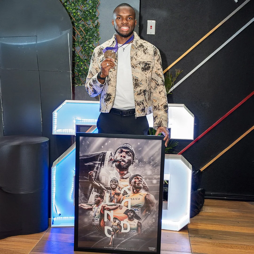 A man holding a gold medal and standing behind a framed poster of athletes, in a modern indoor setting with illuminated geometric decor.