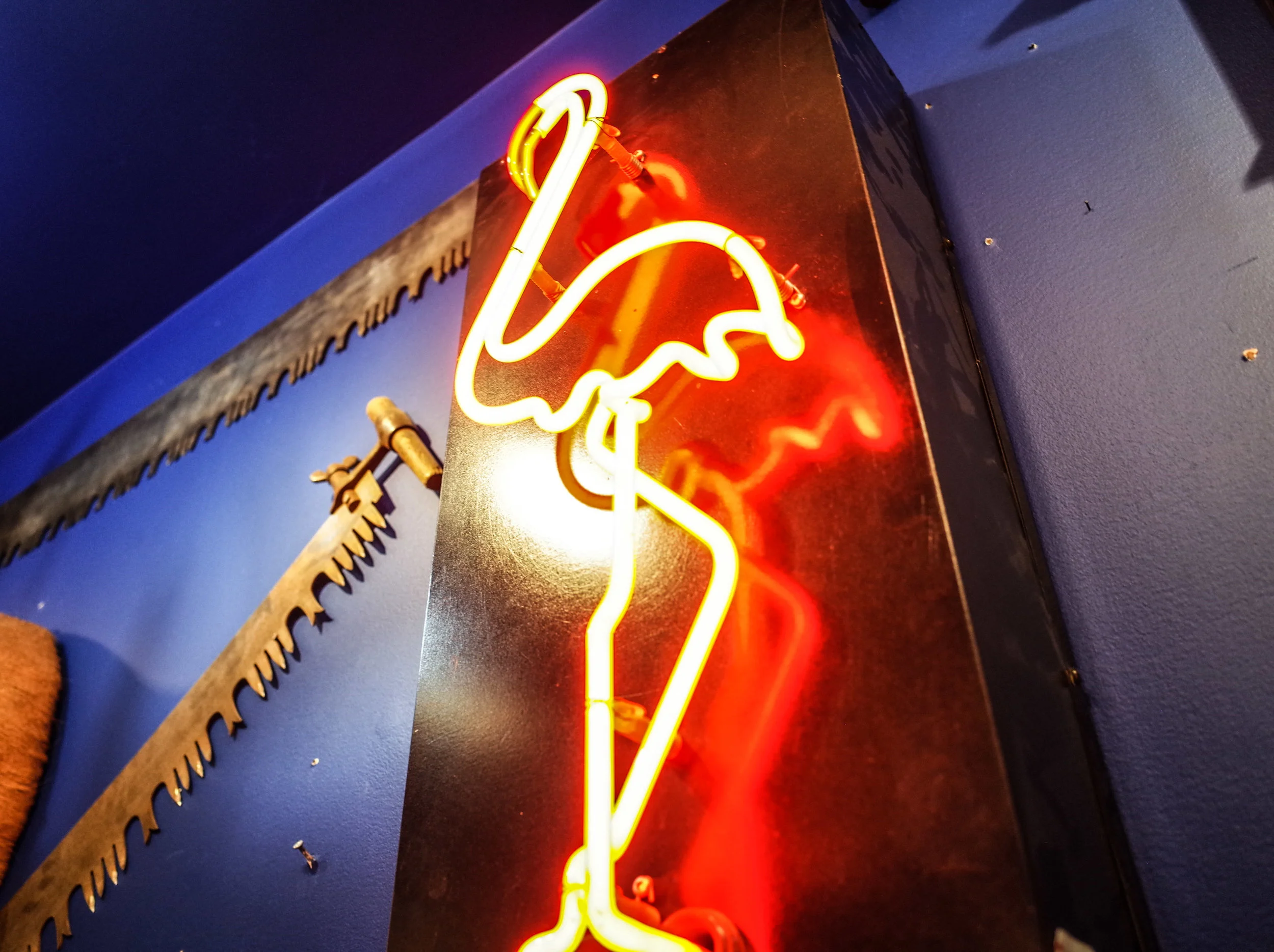 A glowing retro pink flamingo neon sign mounted next to vintage crosscut saws on a blue wall at Space Lab Vancouver, linking to the antique lighting collection.