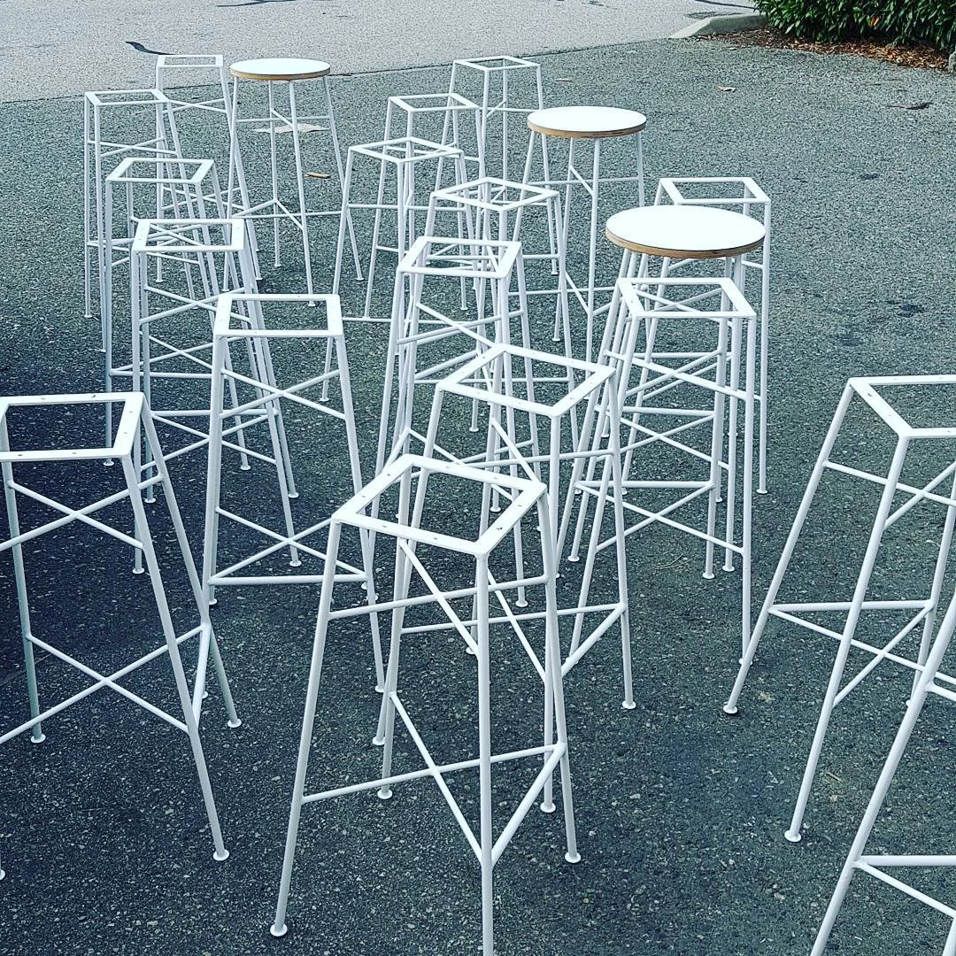 A collection of custom-made white steel bar stool frames on asphalt at Space Lab Vancouver, linking to the custom furniture and welding builds page.