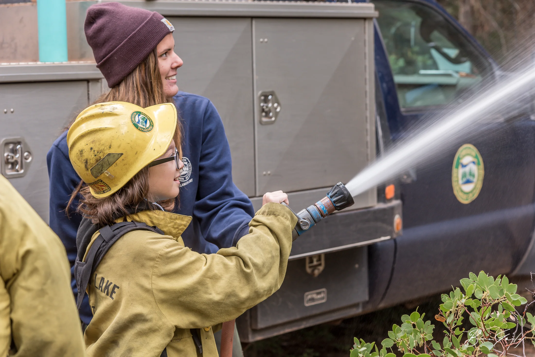 Michelle Cottier a firefighter with ODF geared up Feanix Hightower of Chiloquin Elementary and instructs her how to use the fire hose of Oregon Department of Forestry Engine 8150 at the 54th Annual Klamath County Forest Tour. Image: Brian Gailey
