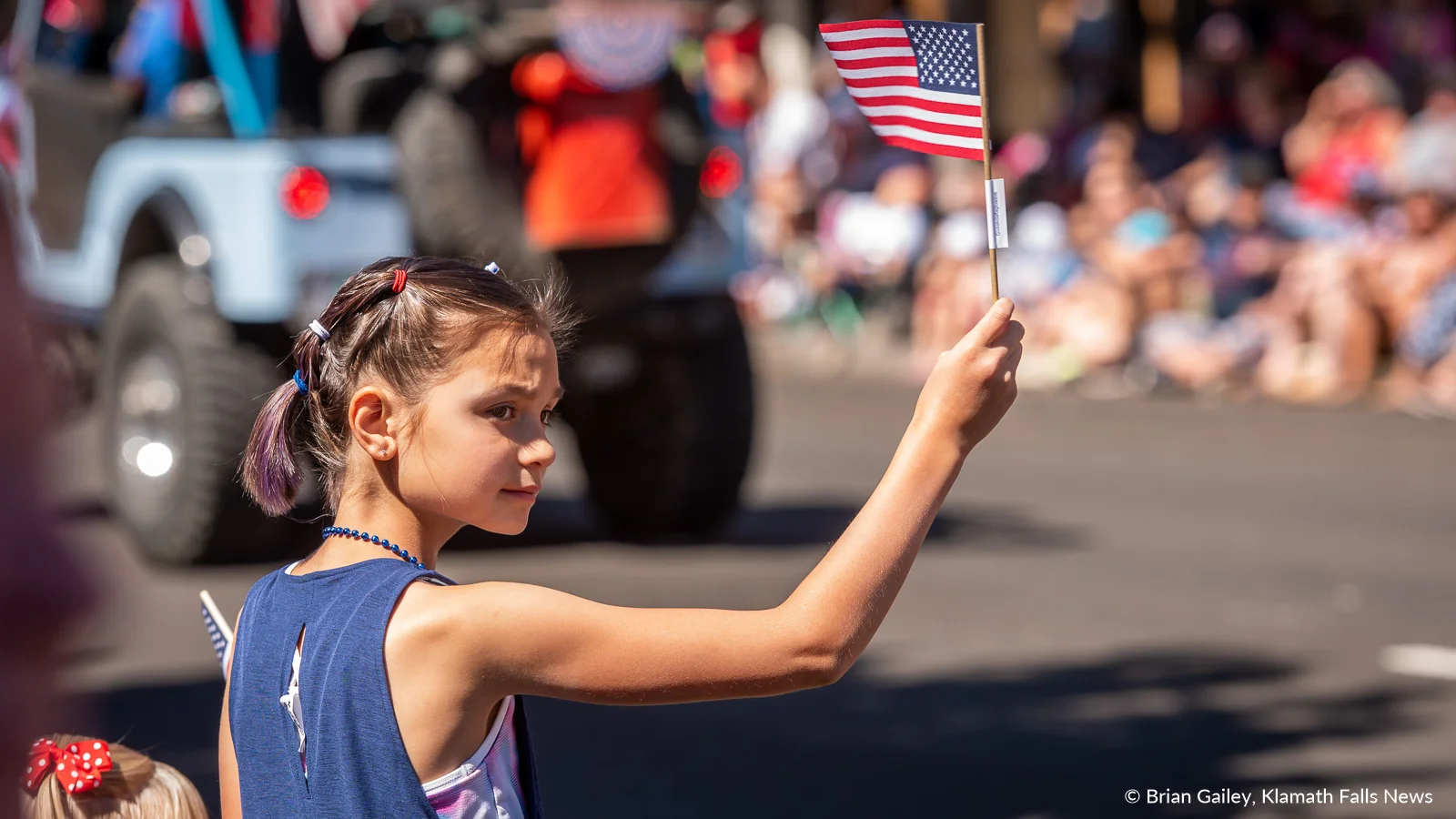 A young lady at the 2019 Klamath Falls Independence Day Parade waves a small US flag given to her. The sticker on it reads, give a kid a flag to wave. July 4, 2019 (Image, Brian Gailey / Klamath Falls News)