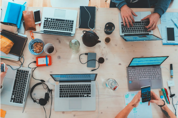 Working too much to feel good enough? Overview shot of laptops and workstations on a wooden desk, with hands working.