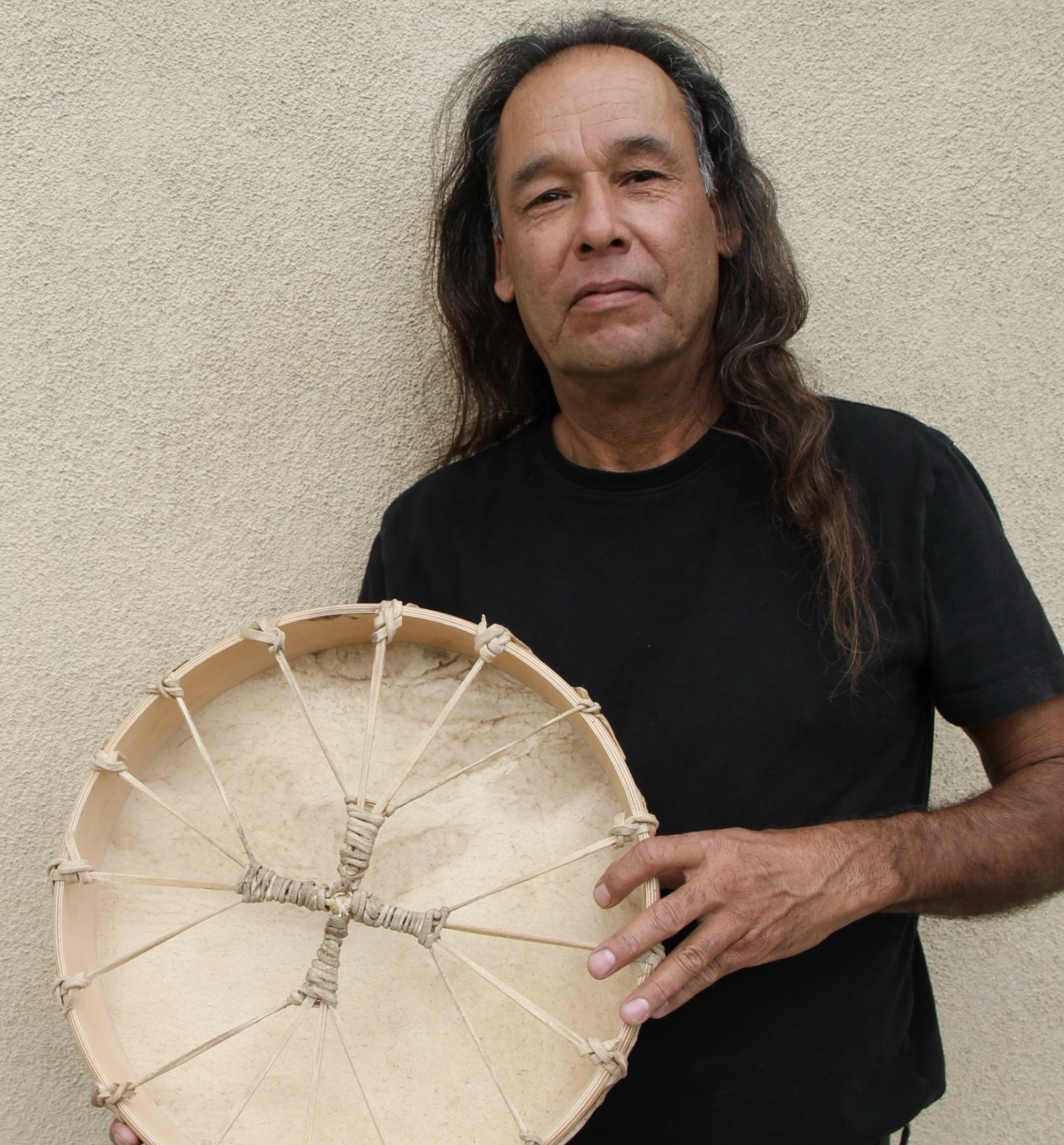 A man with long hair and a black shirt holding a handmade drum with rope details against a beige wall.