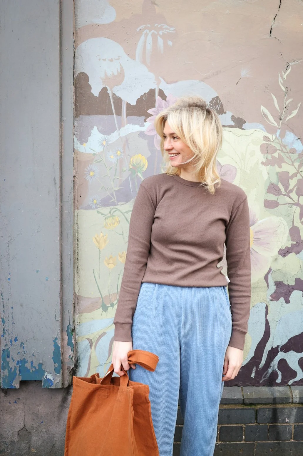 A woman standing outdoors in front of a colorful mural, wearing a mustard yellow t-shirt, striped shorts, holding a woven bag, with blonde hair tied back with a black scrunchie.