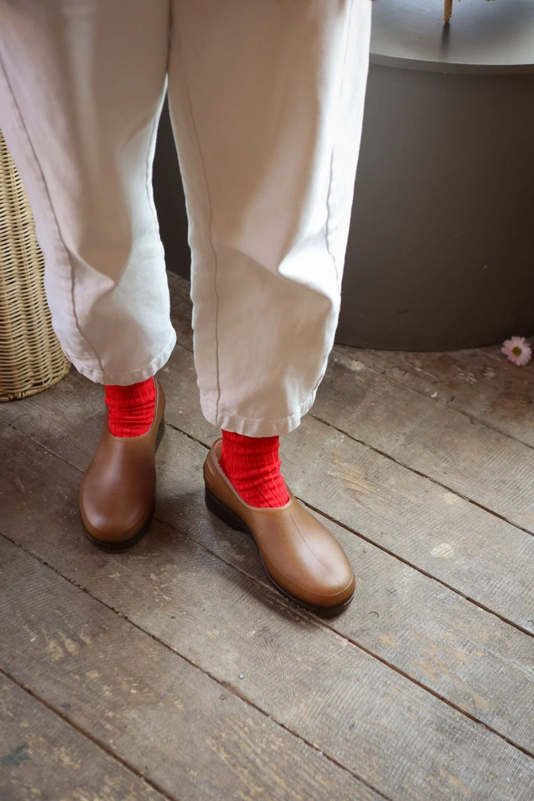 A woman standing outdoors in front of a colorful mural, wearing a mustard yellow t-shirt, striped shorts, holding a woven bag, with blonde hair tied back with a black scrunchie.