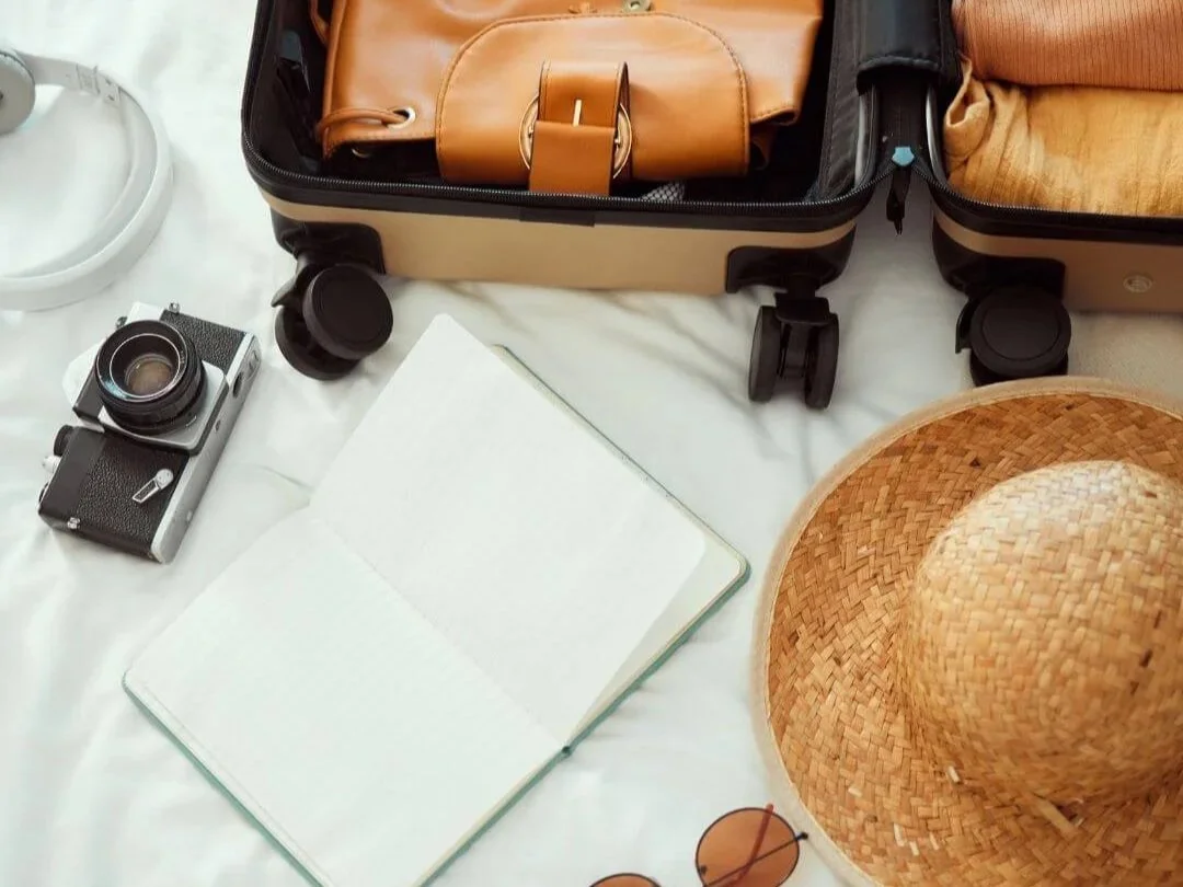 Open suitcase with headphones and a camera, hat and sunglasses on a white sheet. Packing for a trip or vacation.