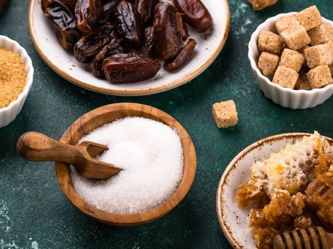 Bowls of sugar, sugar cubes, honey and dates on a green background.