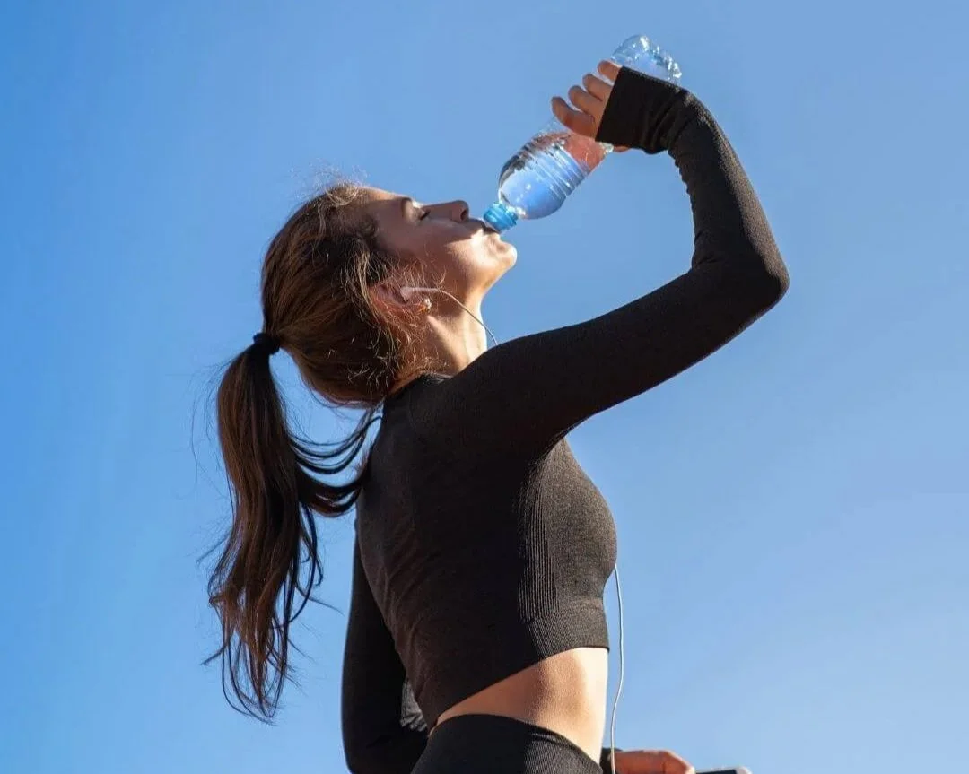 Woman drinking water after a workout on a hot summer day with a blue sky background behind her.