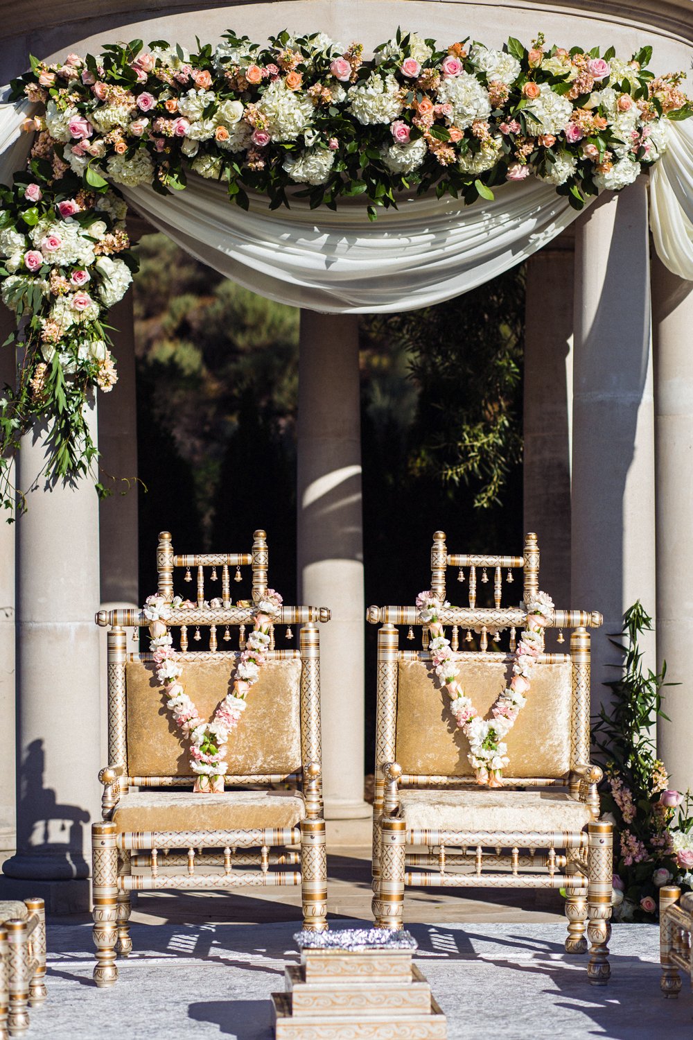 Outdoor wedding altar decorated with pink and orange curtains, floral arrangements, and two ornate chairs on a small elevated stage with stone steps