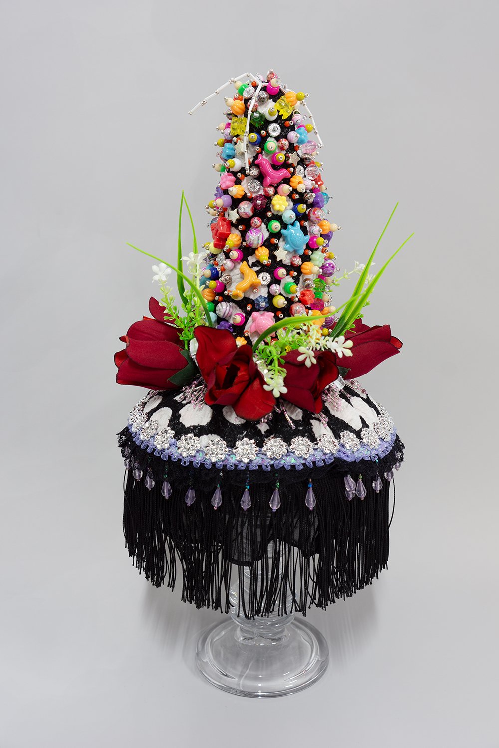   Untitled,  2024, Plastic beads, found fabric, trim, fabric and plastic flowers, buttons, charms, polyester batting, thread, glass stand, 19 x 9 x 9” 