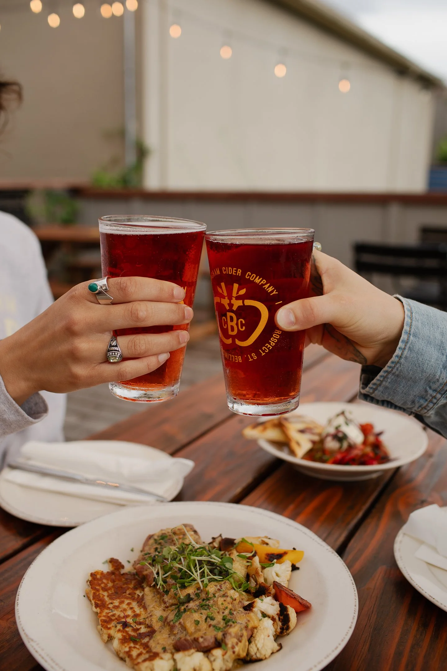 Two people clinking cider over a meal outside of a restaurant