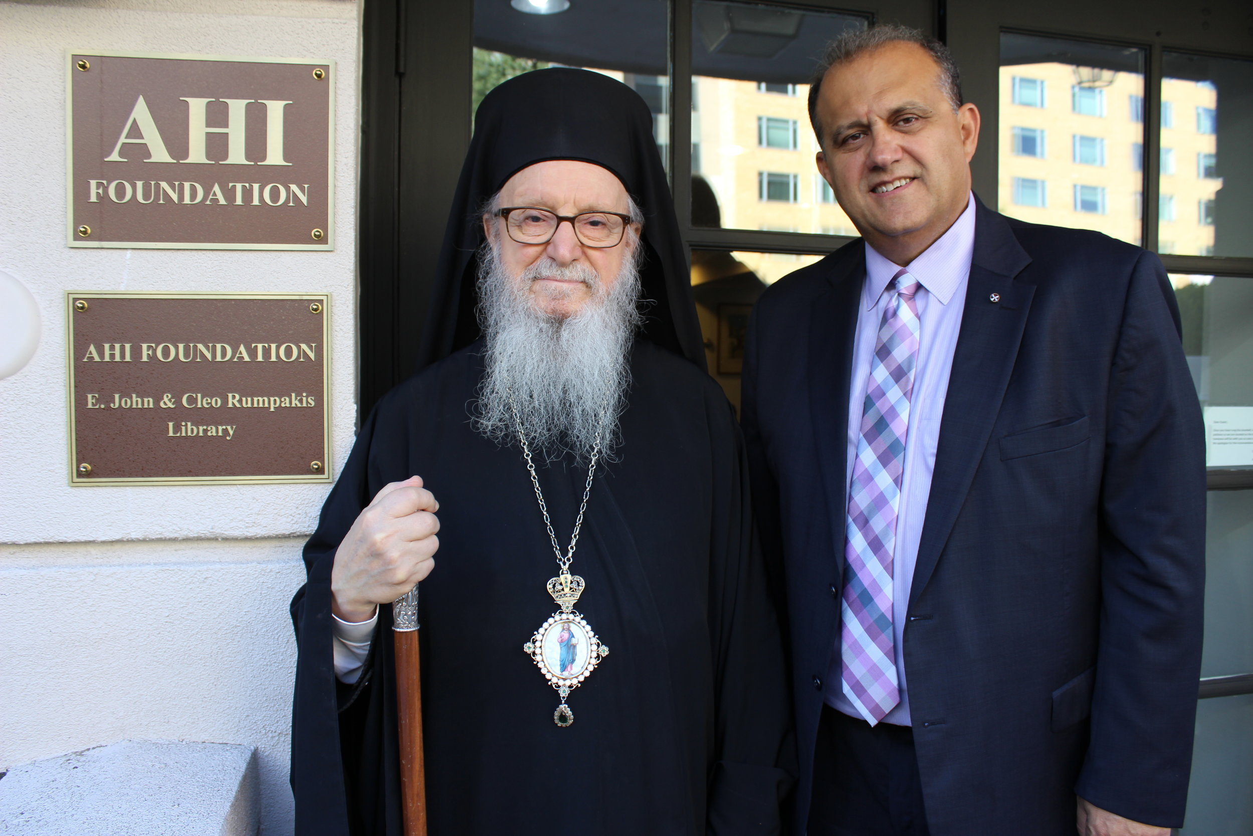 His Eminence Archbishop Demetrios of America and AHI President Nick Larigakis in front of the sign identifying the AHI Foundation E. John &amp; Cleo Rumpakis Library situated at the front entrance to the Hellenic House. 