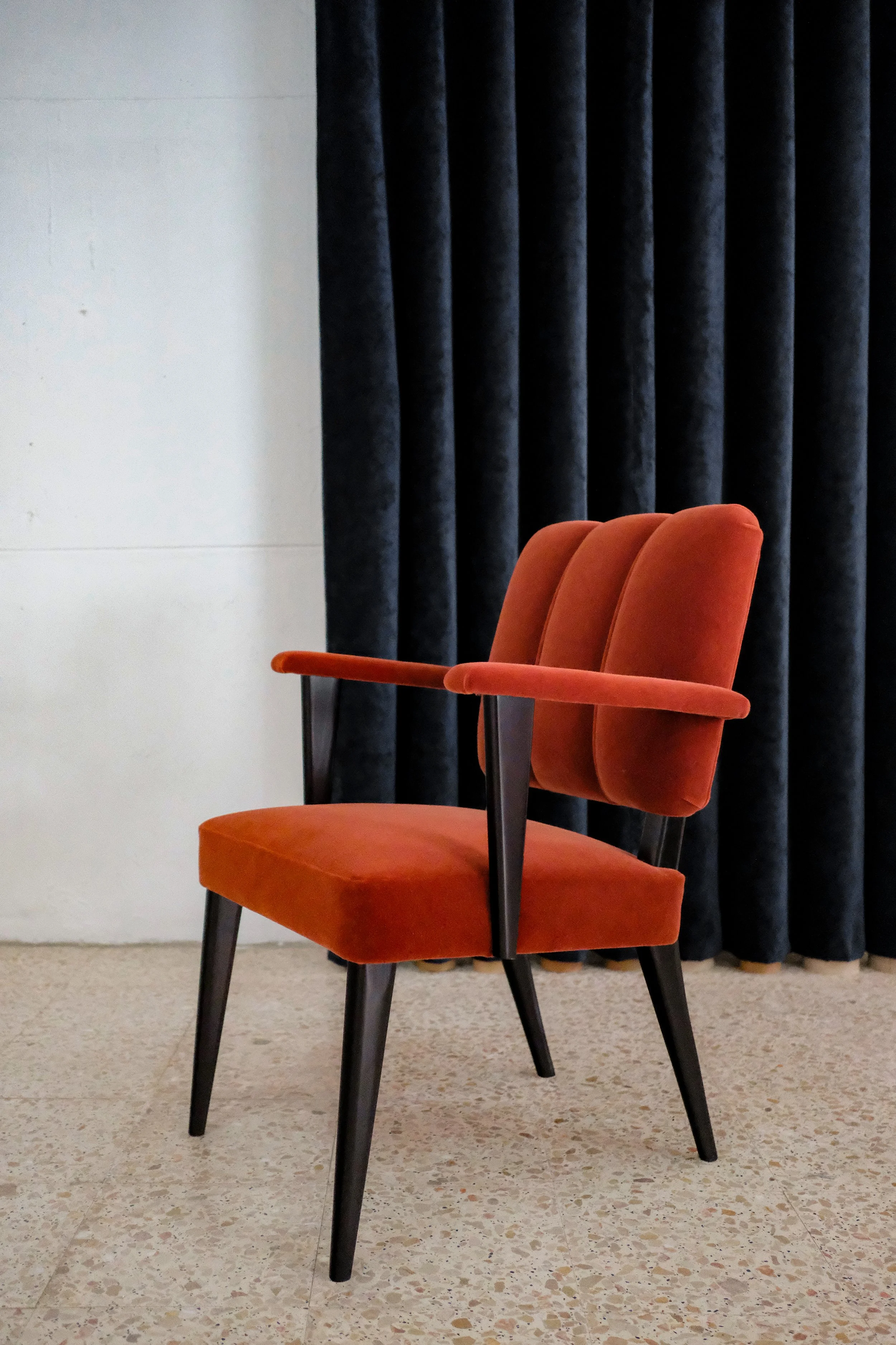 Orange upholstered chair with black legs placed on tiled floor with dark curtain in the background.