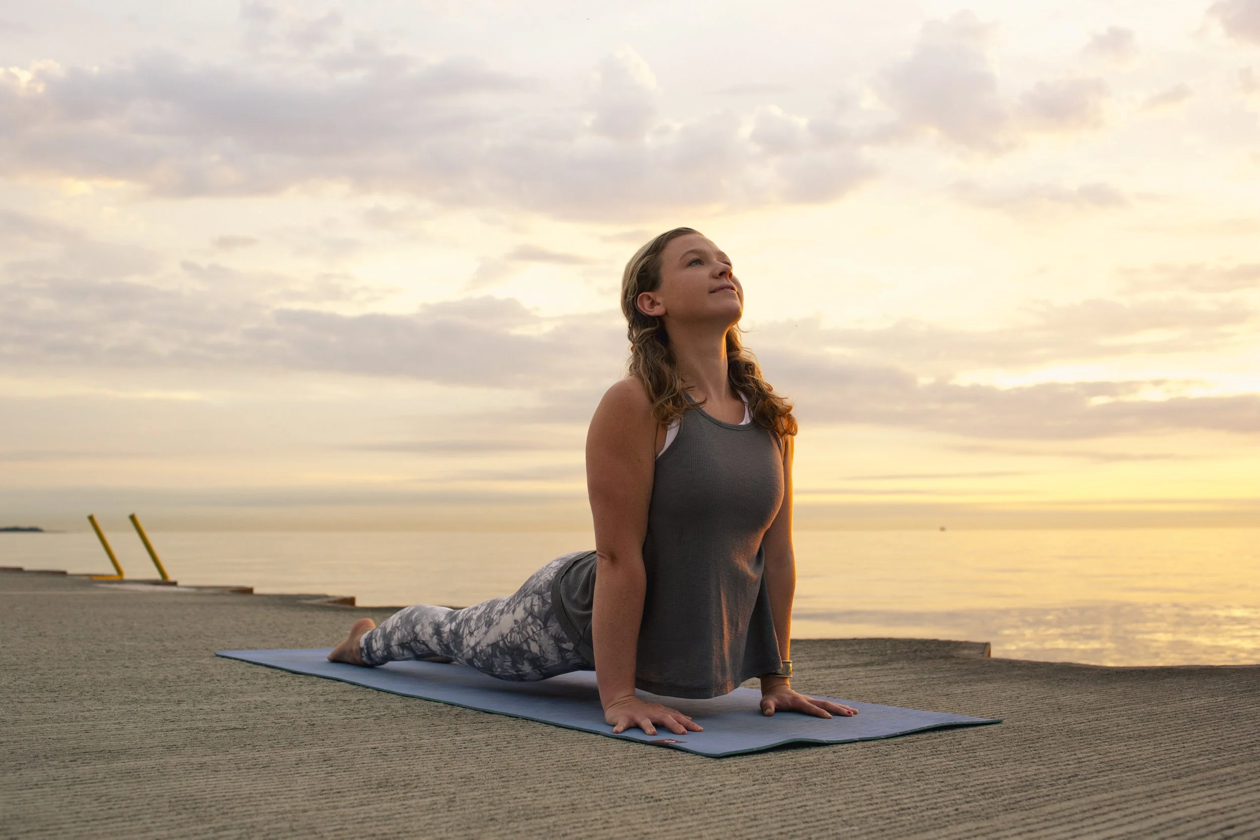 A woman performing a yoga pose called upward dog on a yoga mat outdoors during sunset, with a body of water and a cloudy sky in the background.