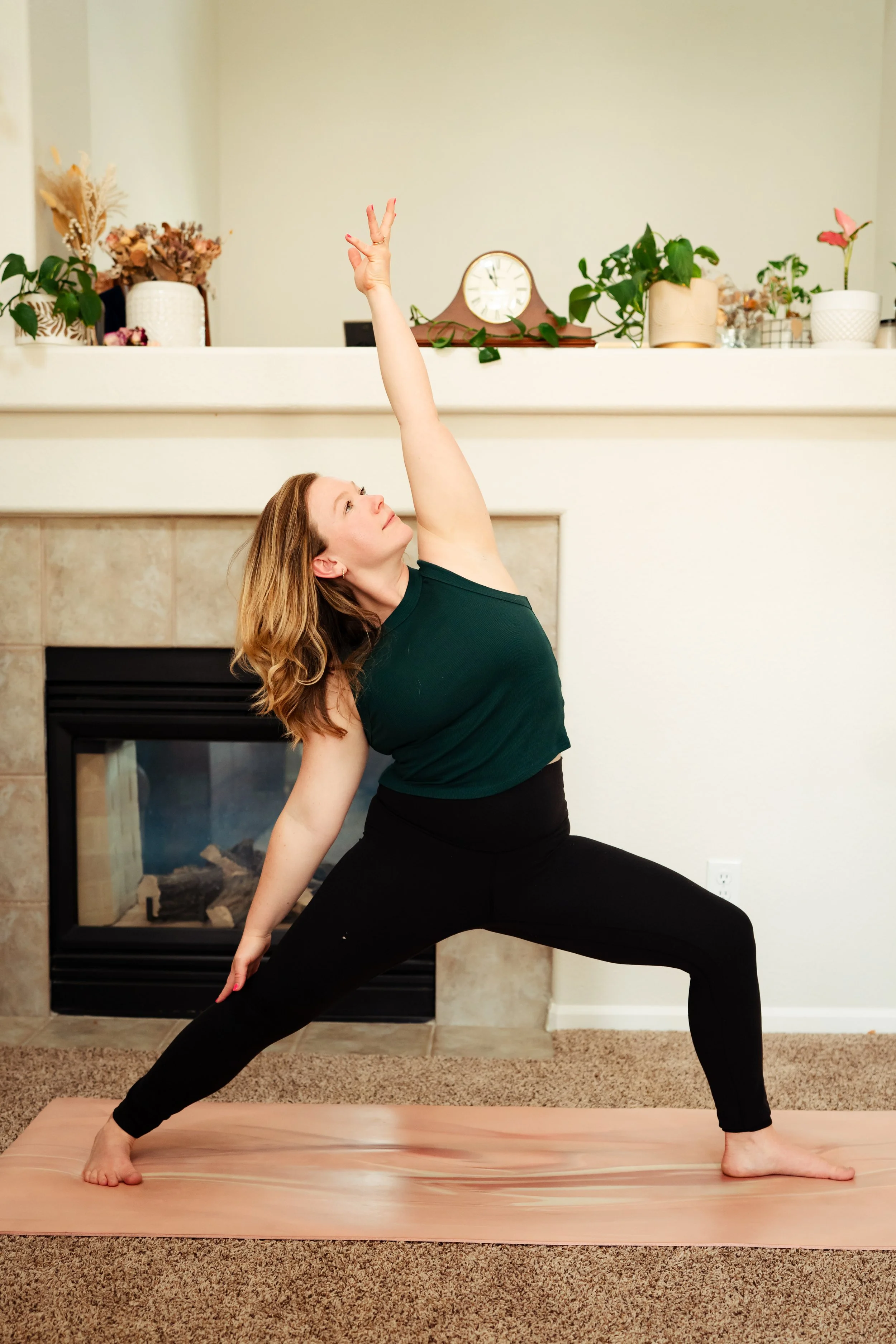Woman doing yoga in lunge pose with one arm raised and the other arm touching the floor, on a yoga mat in a living room with a fireplace and plants.