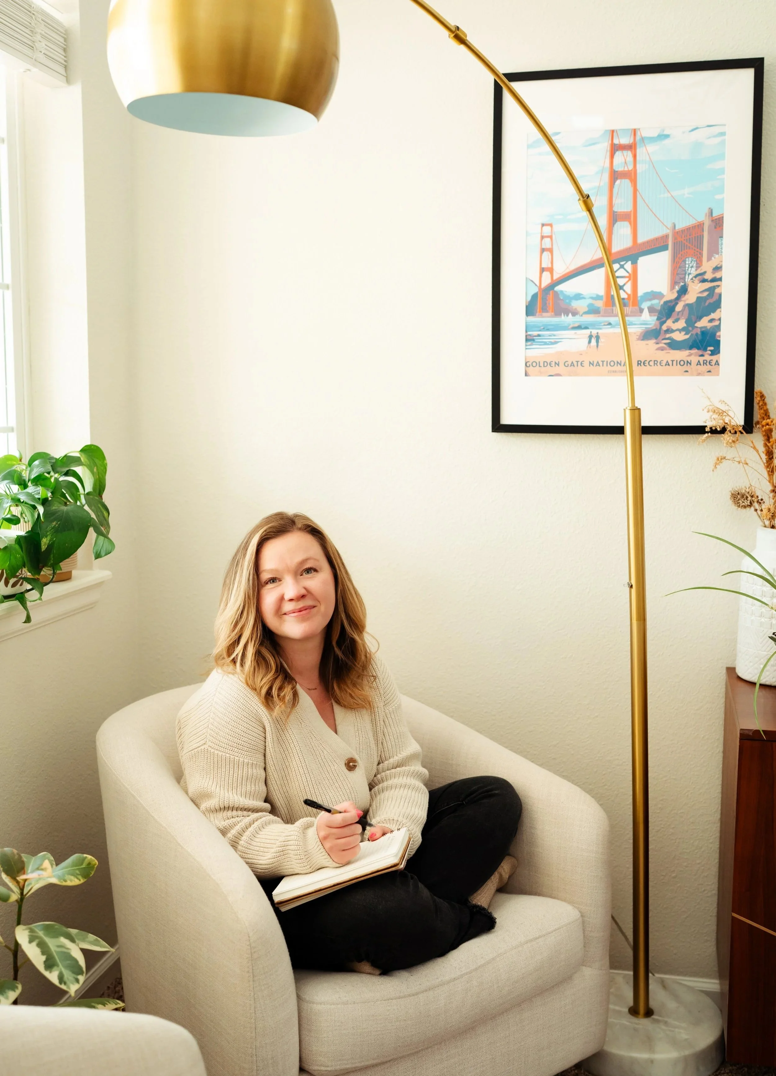 A woman sitting in a light-colored armchair in a room with a framed picture of the Golden Gate Bridge, plants, and a gold arc floor lamp. She is holding a notebook and pen, smiling at the camera.