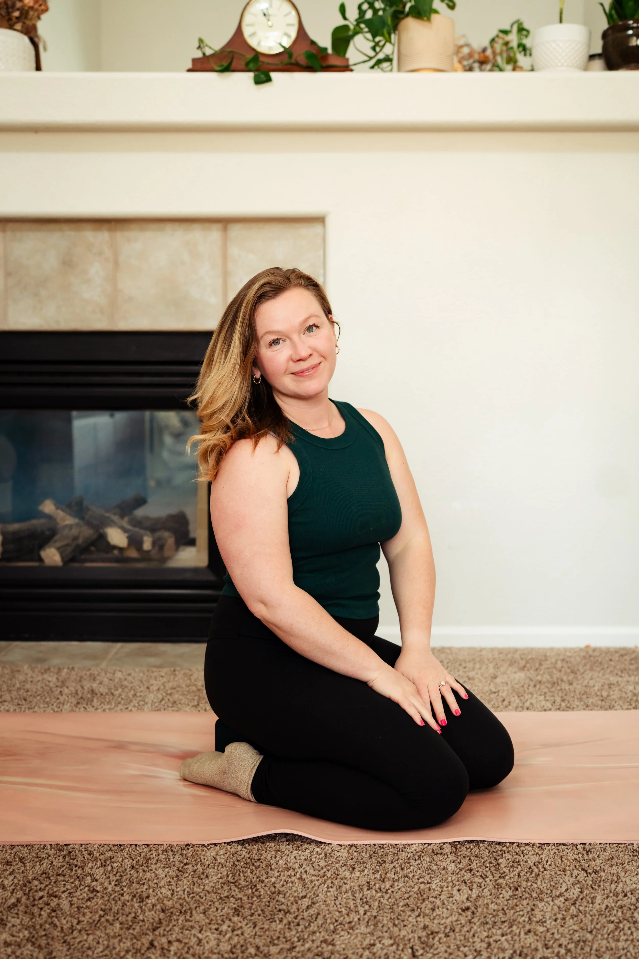 A woman kneeling on a pink exercise mat in front of a fireplace, wearing a dark green sleeveless top and black leggings, with a fireplace and plants in the background.