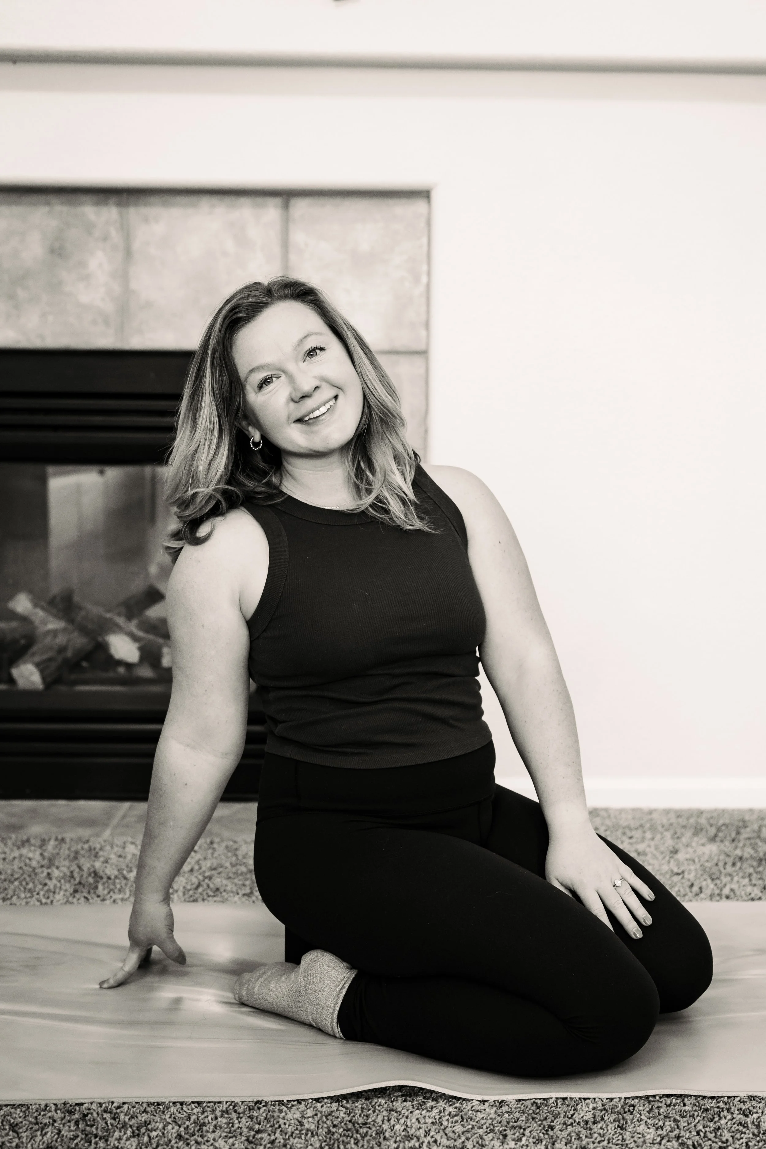 A woman with shoulder-length hair smiling, sitting on a yoga mat next to a fireplace, wearing a sleeveless top and dark pants, in a living room.